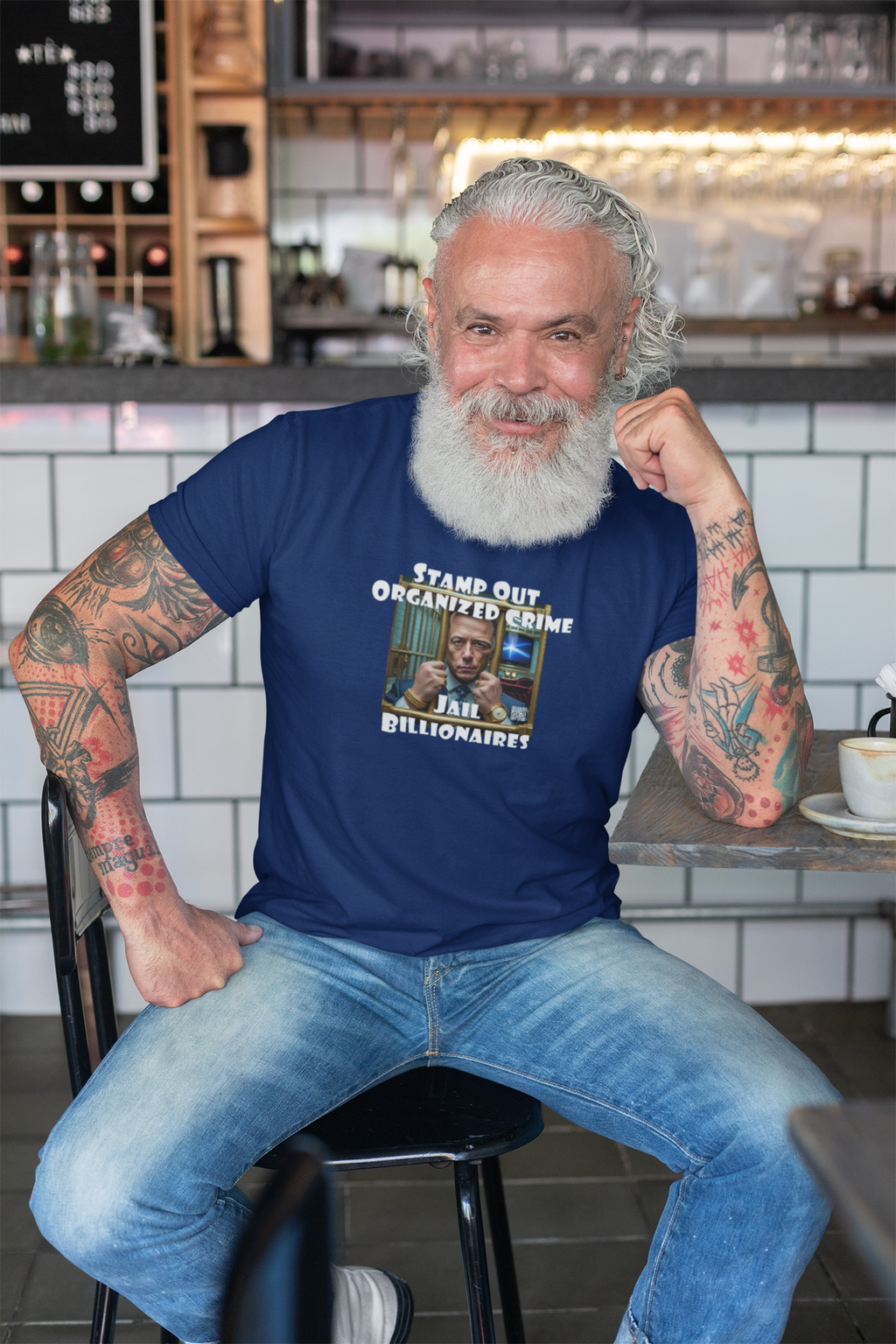 Man with a beard wearing a Stamp Out Organized Crime-Jail Billionaires Activist T-shirt in a café setting.