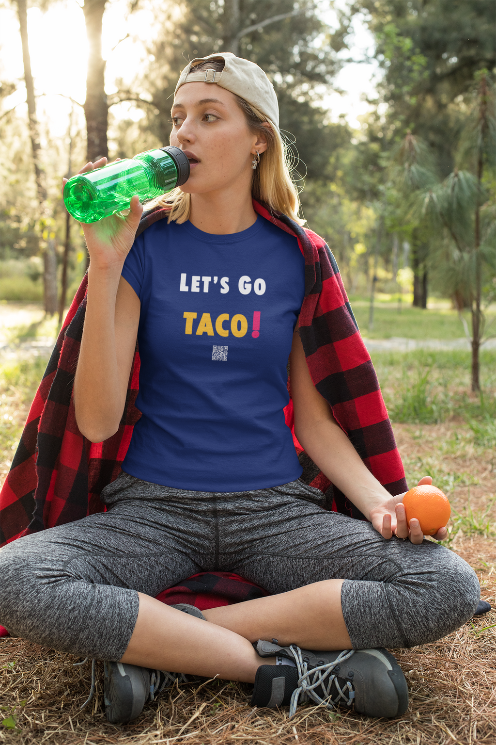 Woman wearing a TACO Activist TShirt, holding a green water bottle and orange, sitting outdoors in nature.