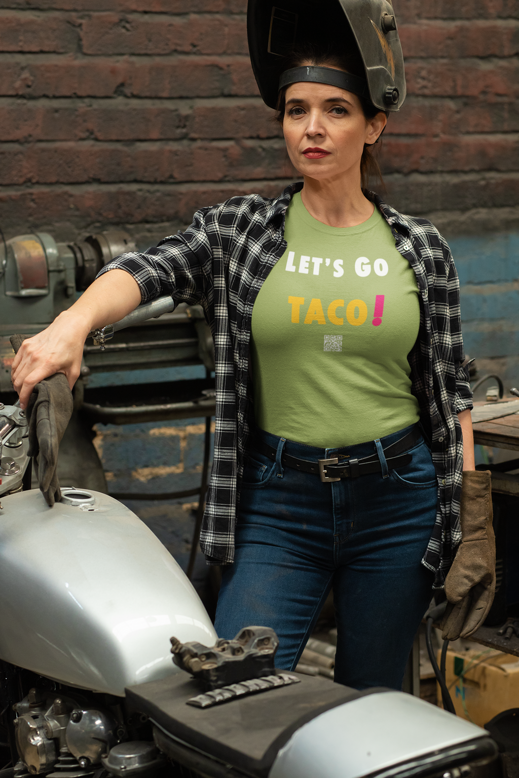 Woman wearing a TACO Activist TShirt in a workshop, showcasing a confident pose next to a motorcycle.