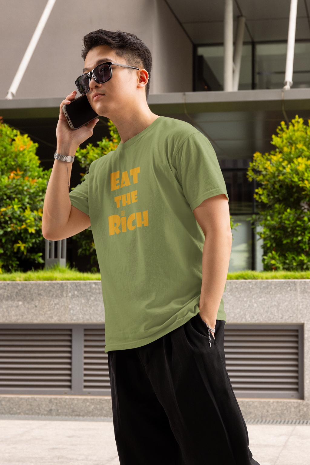 A person wearing an EAT THE RICH Unisex TShirt in an urban setting, showcasing bold text design.