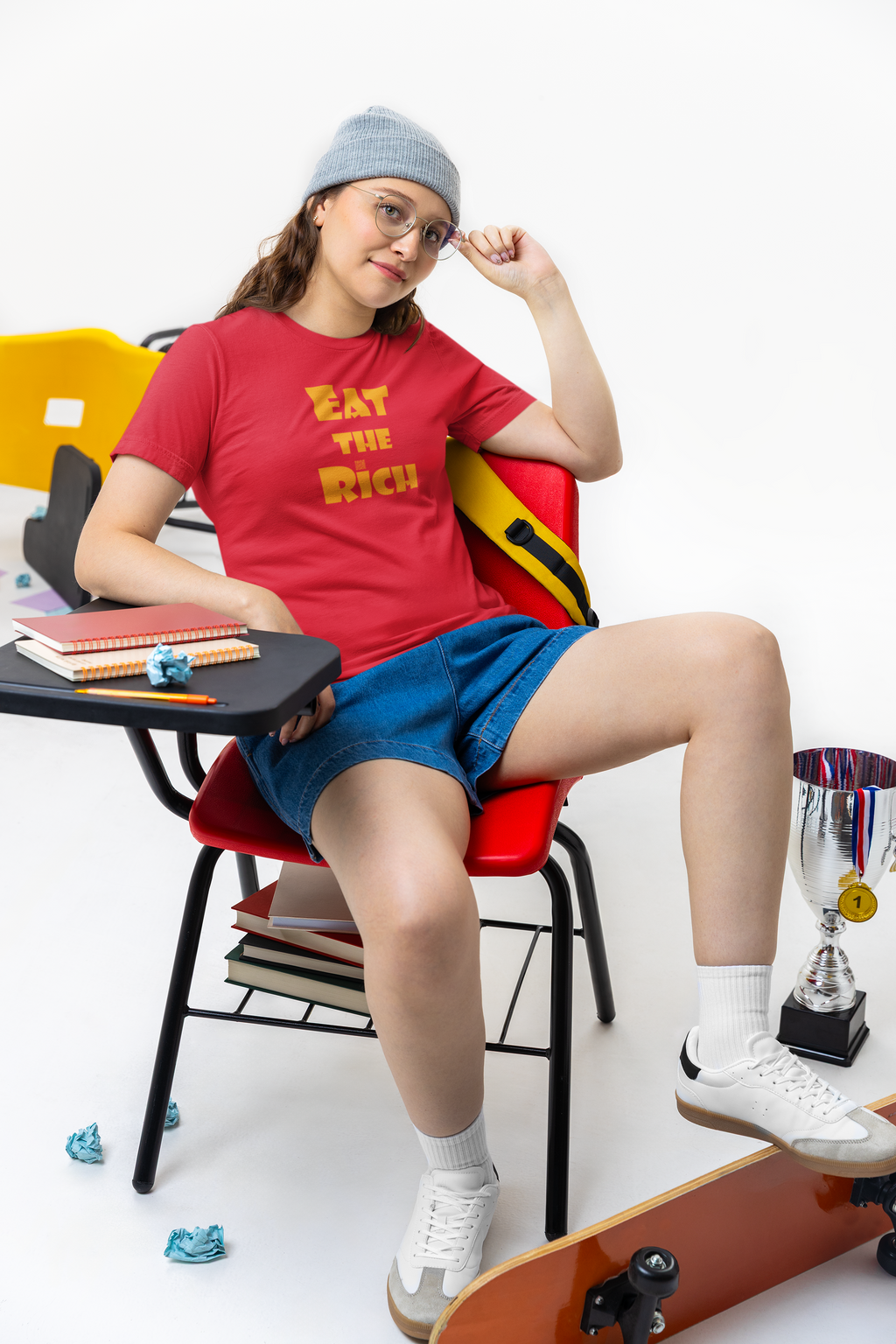 A person wearing an EAT THE RICH Unisex TShirt, sitting on a chair with school supplies and a skateboard.