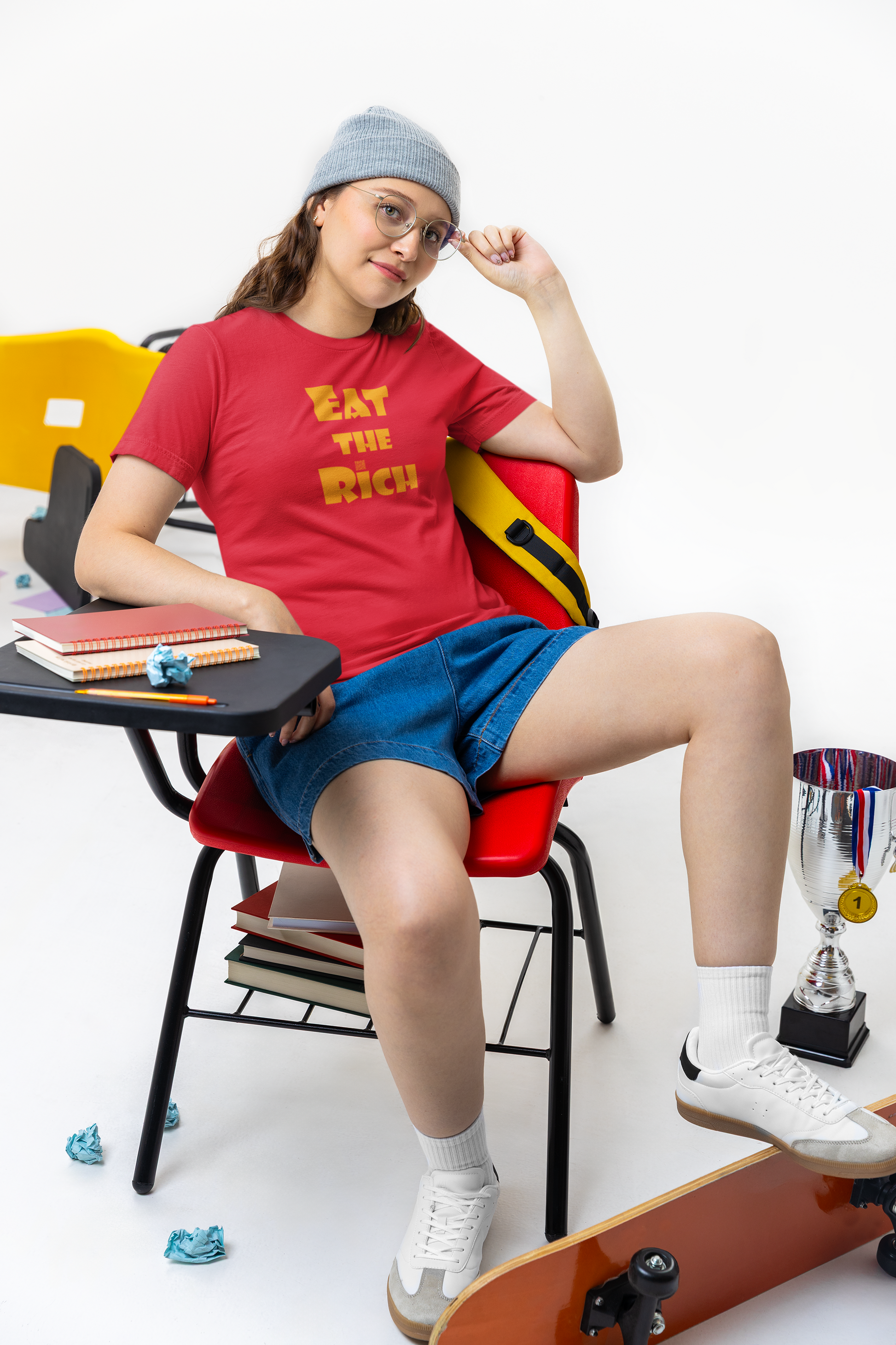 A person wearing an EAT THE RICH Unisex TShirt, sitting on a chair with school supplies and a skateboard.