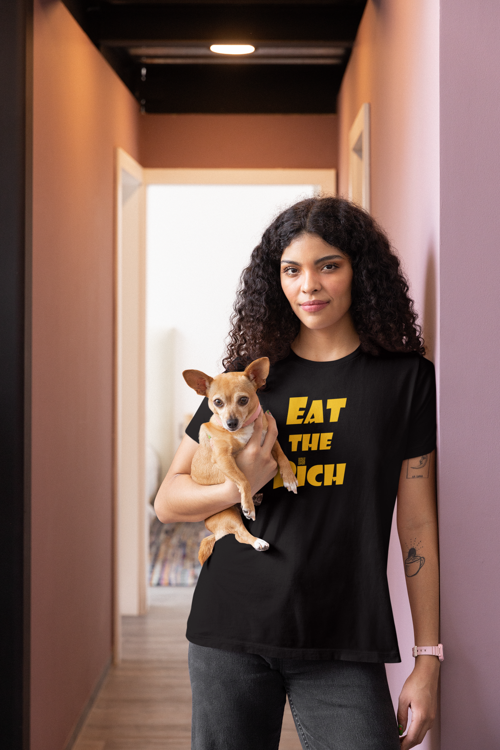 Model wearing EAT THE RICH Unisex TShirt while holding a small dog in a modern hallway.
