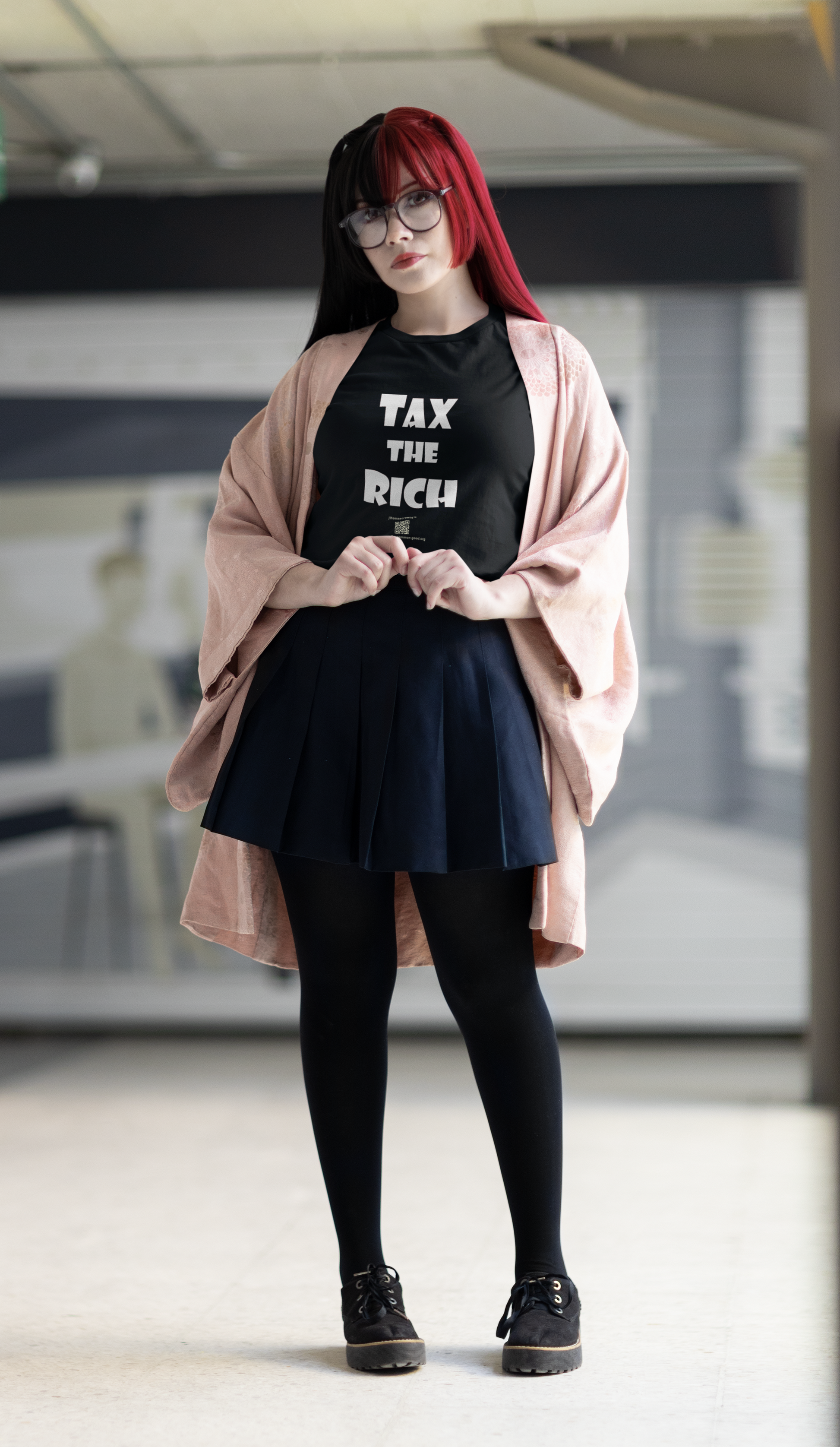 A model wearing a TAX THE RICH Unisex T-Shirt with a pink cardigan and navy skirt, standing in a modern setting.