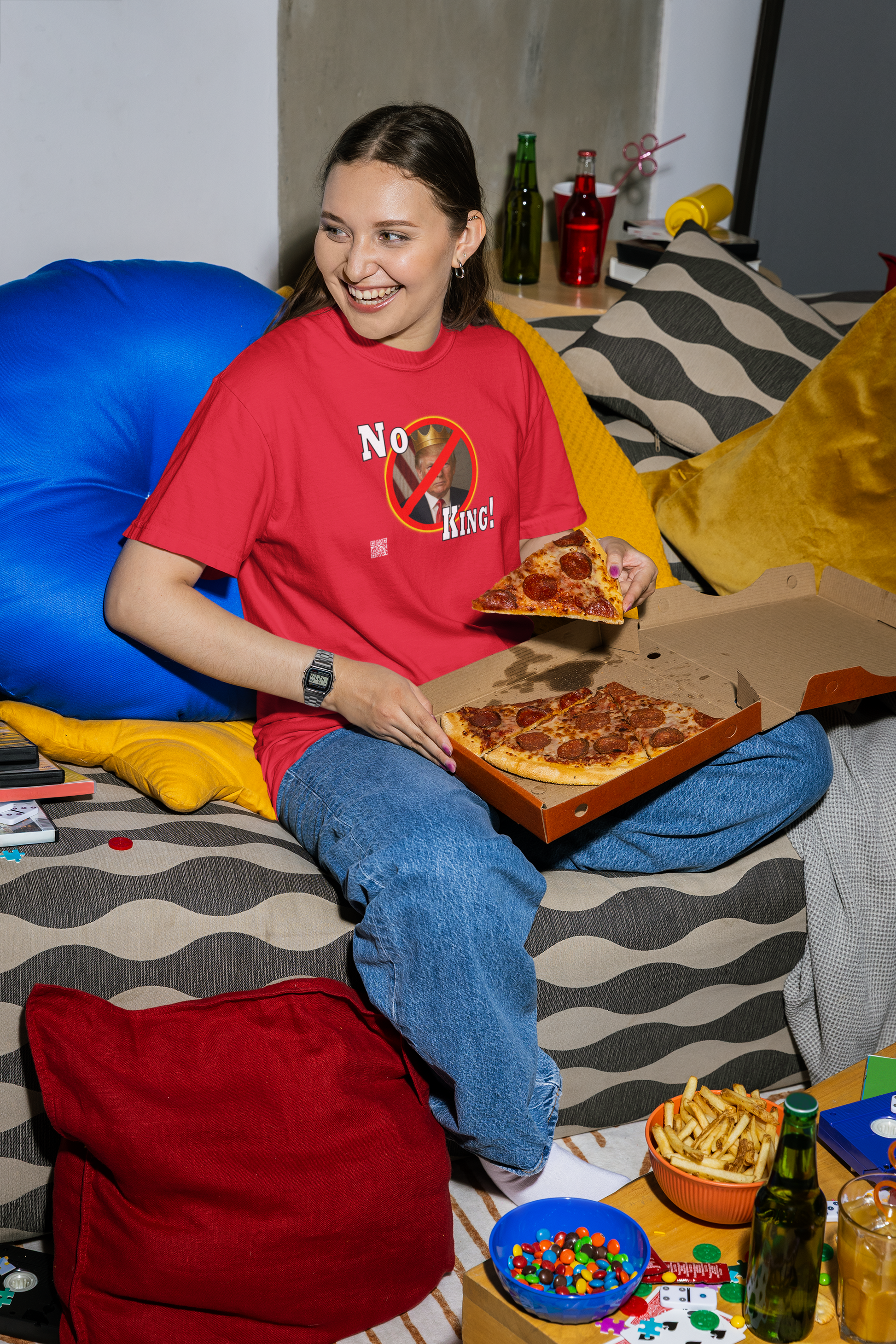 Smiling woman in a No King Activist TShirt holding pizza, surrounded by snacks in a cozy living room setting.