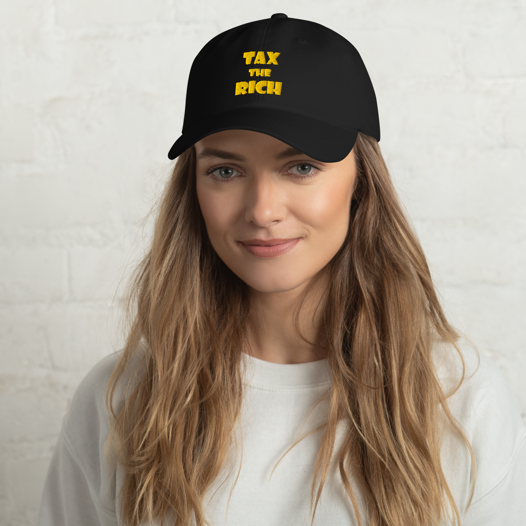 TAX THE RICH — Embroidered Dad Hat worn by a woman with long hair against a white brick wall background.