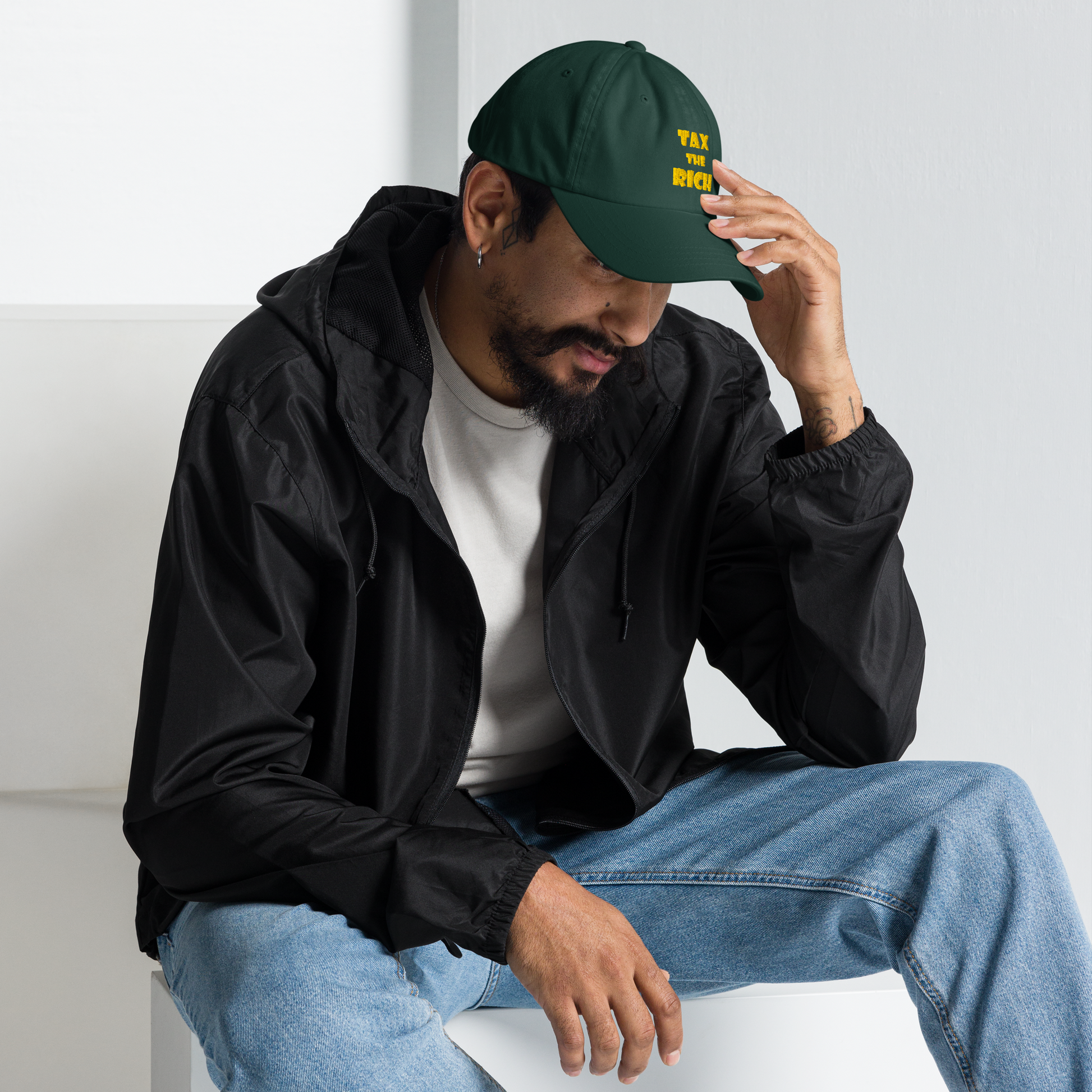 Man wearing a TAX THE RICH — Embroidered Dad Hat and black jacket, sitting and looking thoughtful.