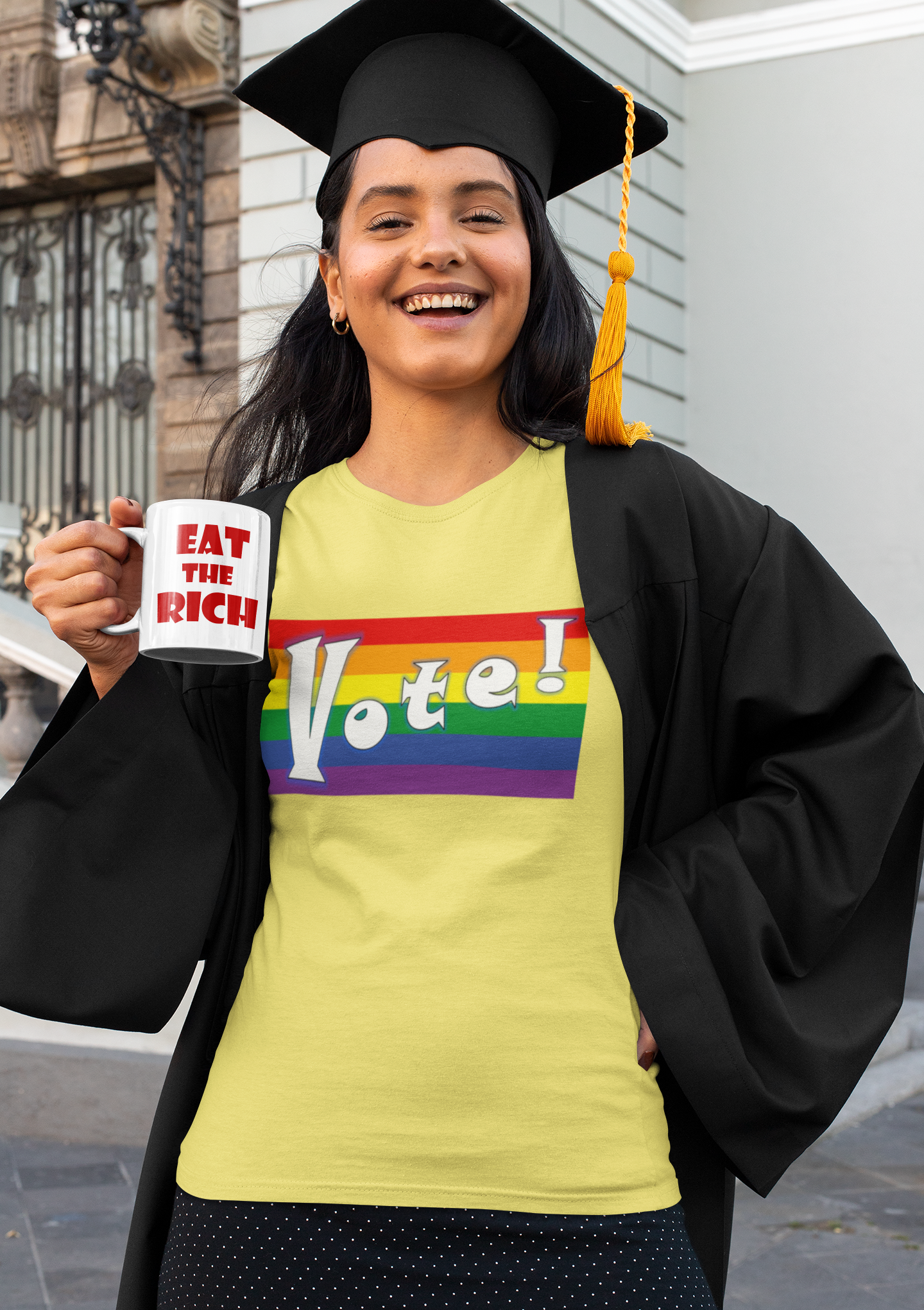 Smiling graduate holding a White EAT THE RICH 11oz Ceramic Mug and wearing a Vote! rainbow shirt.