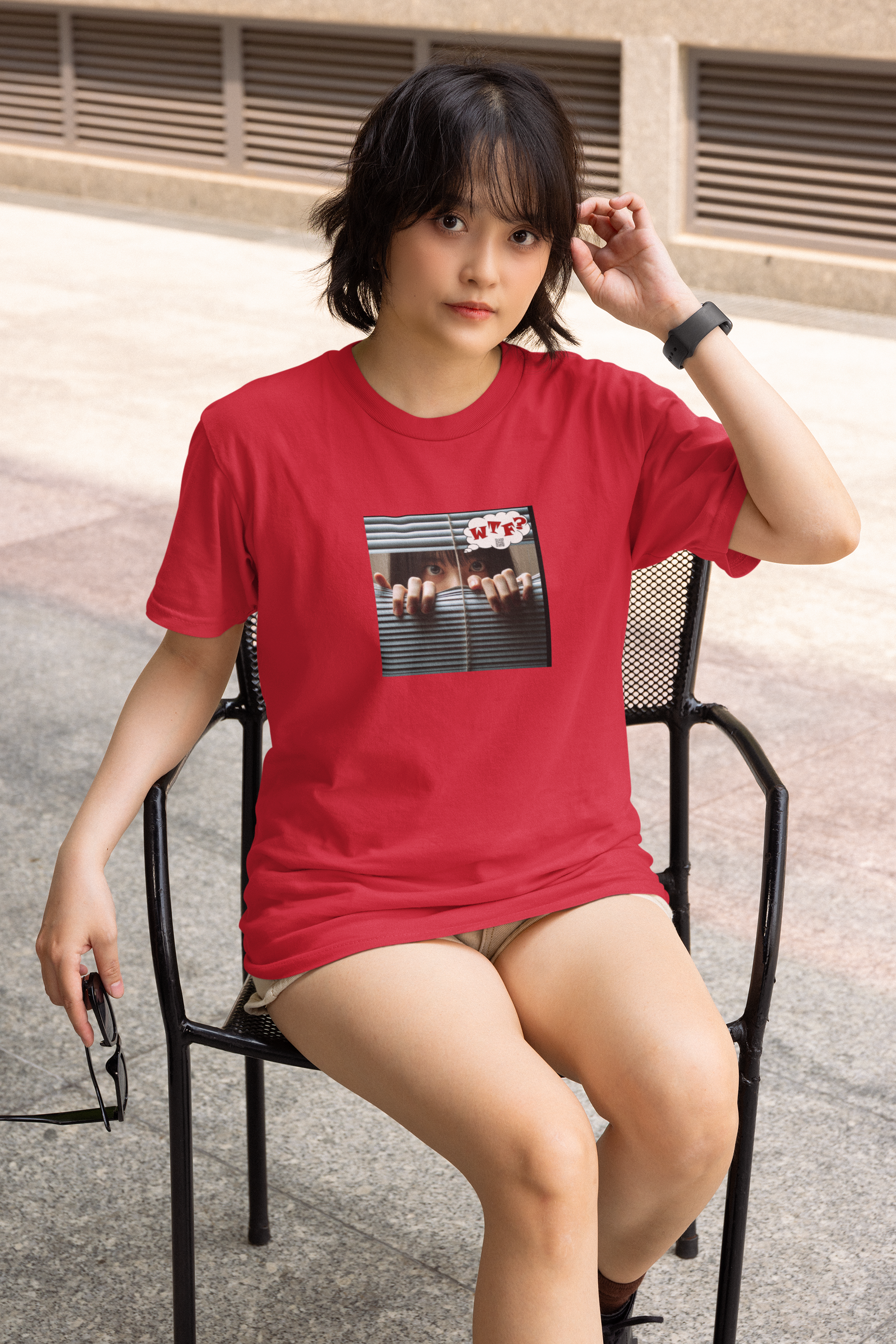 asian woman sitting in a black chair on a sidewalk while wearing shorts and a cardinal red WTF? protest tshirt featuring an asian woman holding slats of a venetian blind apart while she is thinking WTF? in red lettering
