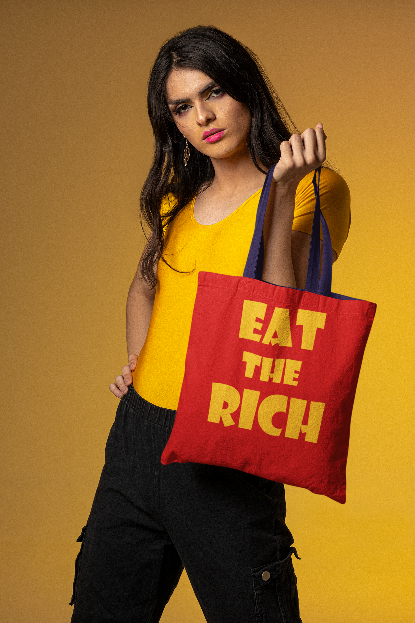 A model holding an Eat the Rich accessory tote bag with bold typography against a yellow background.