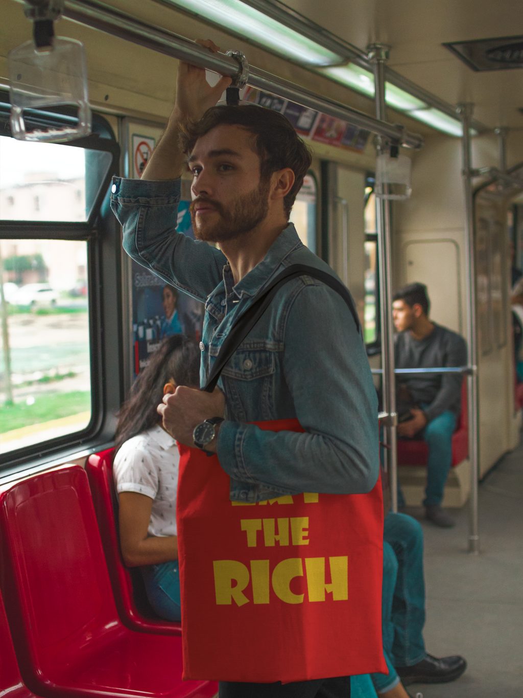 Man on public transport holding Eat the Rich accessory tote bag with bold typography.