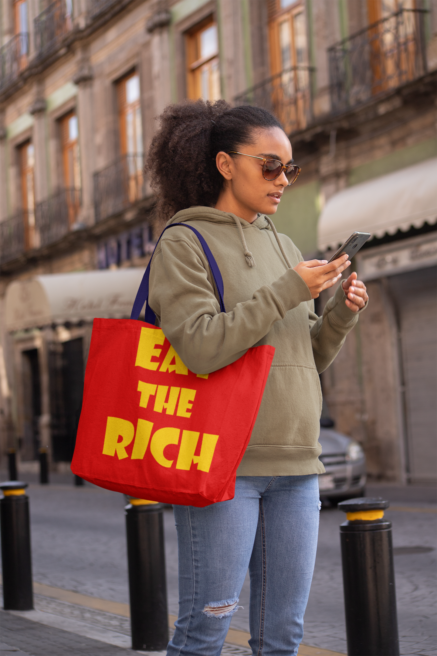 Young woman in street holding smartphone with Eat the Rich accessory tote bag.