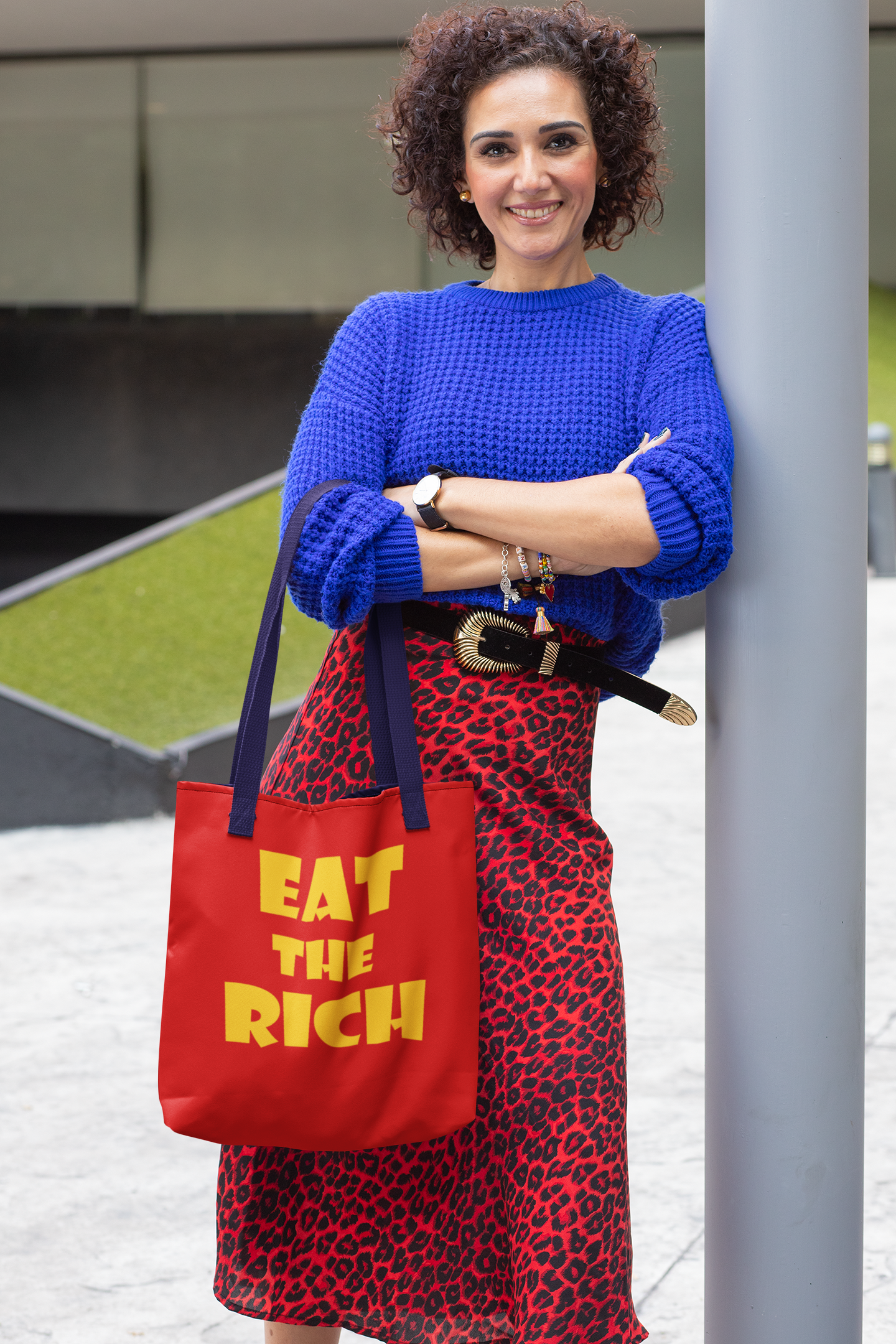 Model wearing a blue sweater and red leopard print skirt, holding an Eat the Rich accessory tote bag with bold typography.