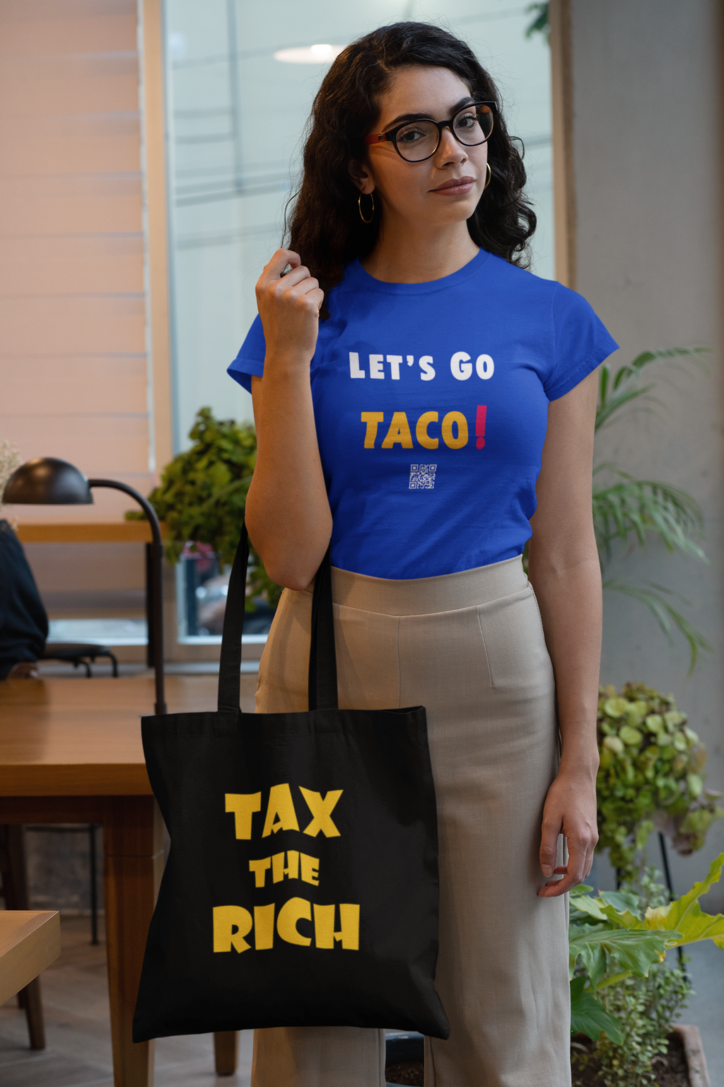 A woman wearing a blue shirt and beige pants holding a Tax the Rich accessory tote bag in a modern indoor setting.
