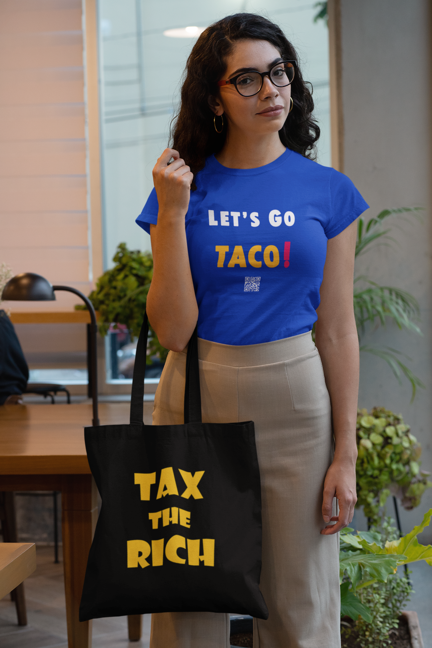 A woman wearing a blue shirt and beige pants holding a Tax the Rich accessory tote bag in a modern indoor setting.