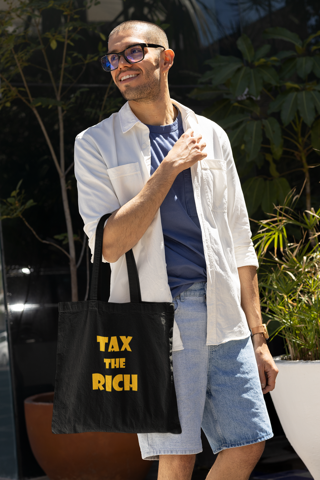 A person smiling while holding a Tax the Rich accessory tote bag in a sunny outdoor setting.