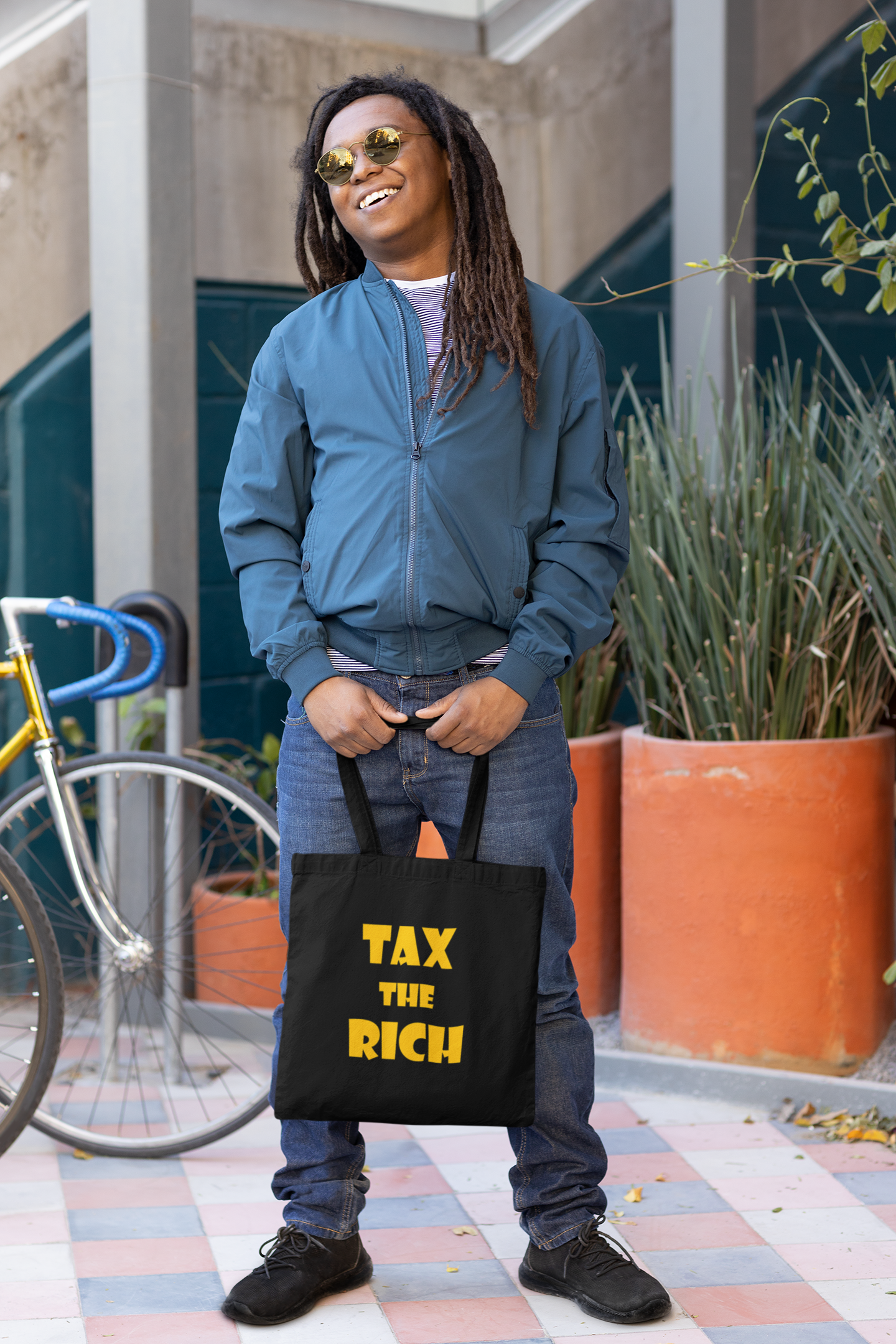 Person holding a Tax the Rich accessory tote bag with bold text, standing next to a bicycle and greenery.