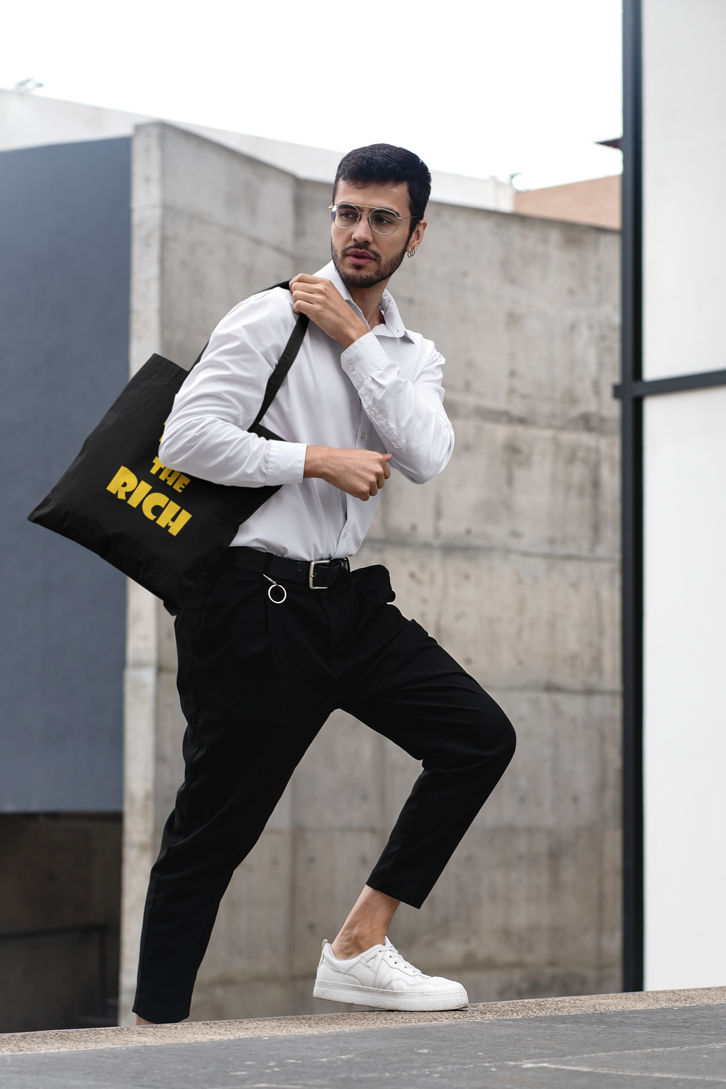 Man wearing white shirt and black pants carrying a Tax the Rich accessory tote bag in an urban setting.