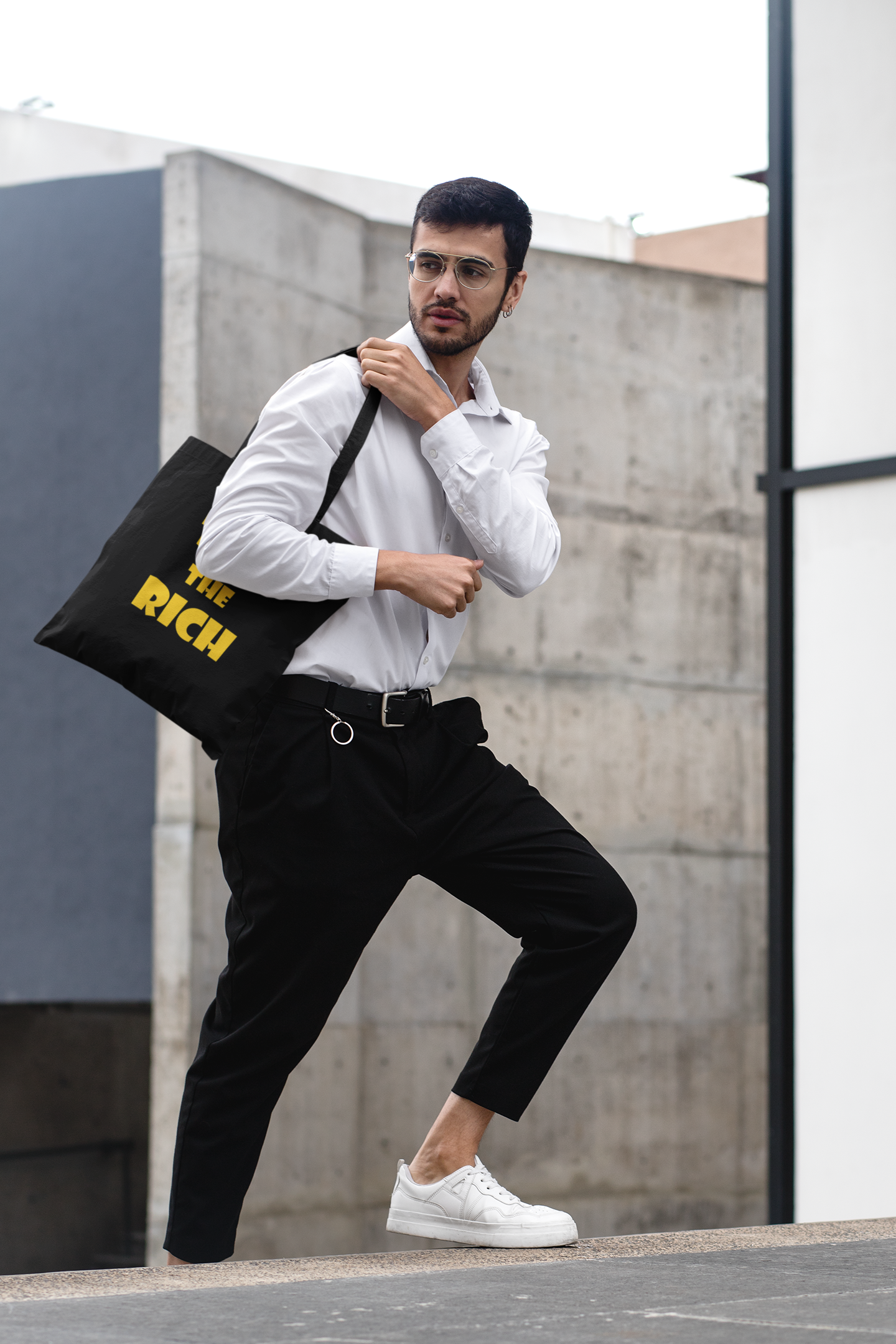 Man wearing white shirt and black pants carrying a Tax the Rich accessory tote bag in an urban setting.