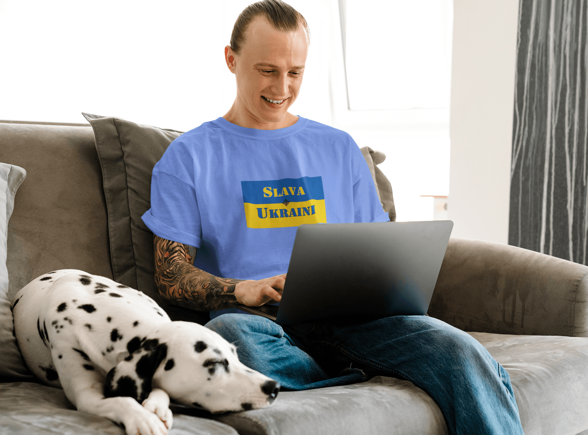 Person wearing a Slava Ukraini Tee Shirt while working on a laptop, with a dog resting nearby.