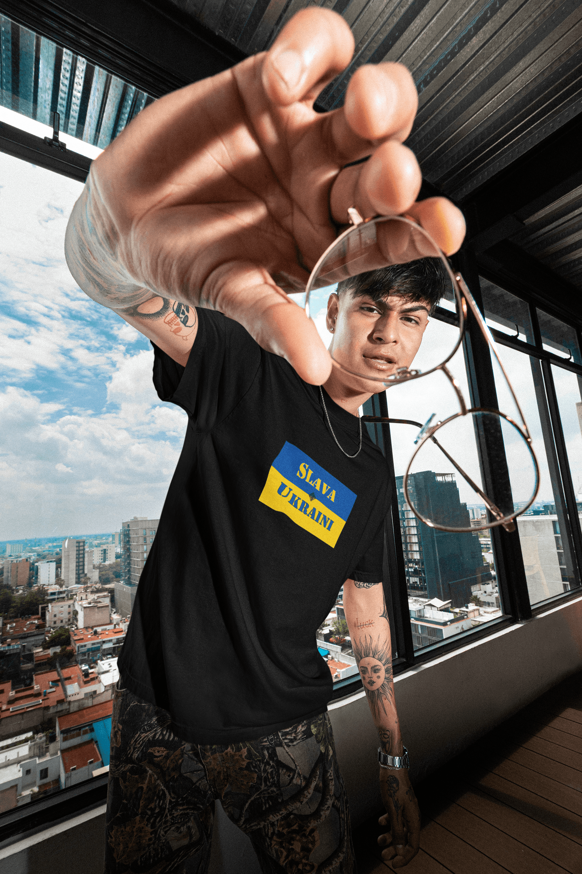 Young Hispanic man wearing an activist black Slava Ukraini Tshirt standing in front of a picture window overlooking the city