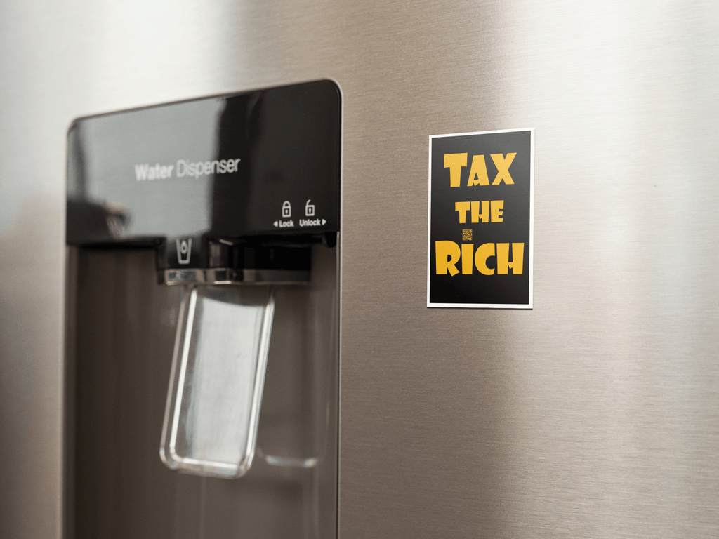 4" x 5" black vinyl magnet with Tax the Rich in gold lettering stuck to the door of a stainless steel refrigerator door