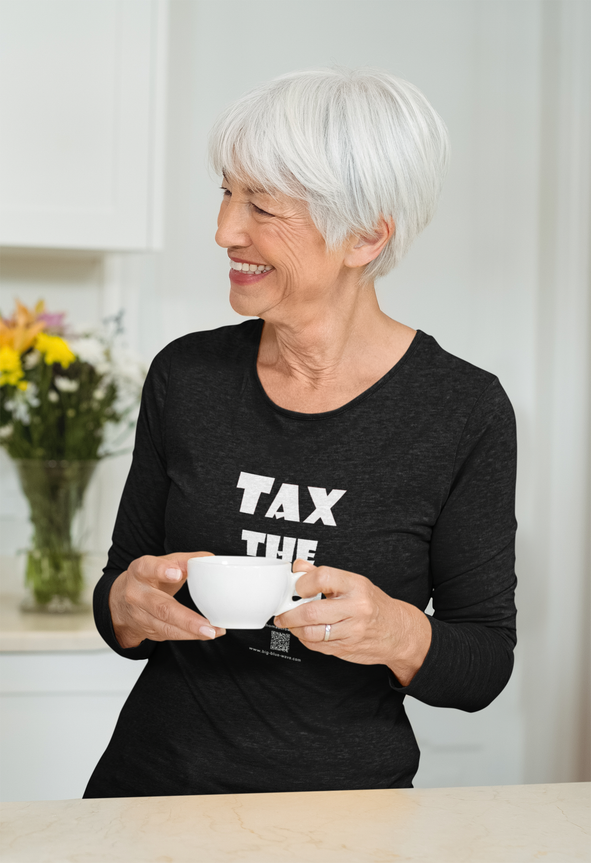An older woman with short white hair is smiling and holding a white mug in a bright kitchen setting. She is wearing a dark charcoal heather long-sleeve t-shirt with the words 'TAX THE' printed in bold white capital letters on the front.