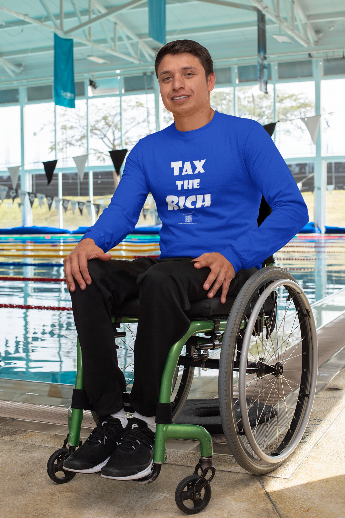 A man sitting in a green manual wheelchair by an indoor swimming pool, wearing a bright blue long-sleeve t-shirt with the white text 'TAX THE RICH' on the front.
