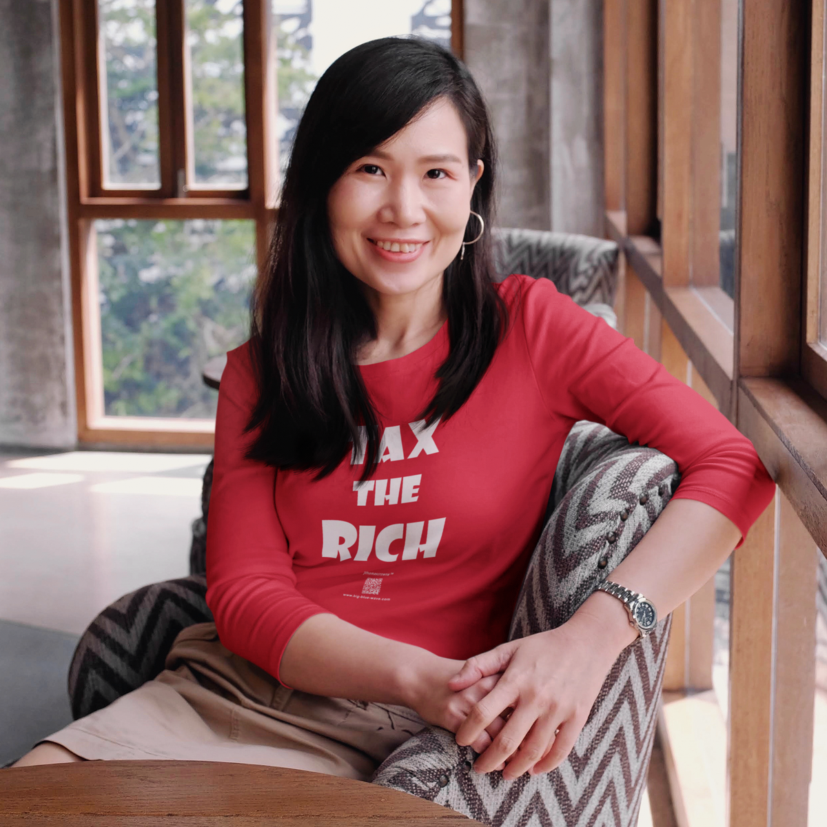 A woman sitting in a patterned armchair, wearing a red long-sleeve t-shirt with the text 'MAX THE RICH' in white. The photo is taken in a warm, sunlit room with wooden window frames.