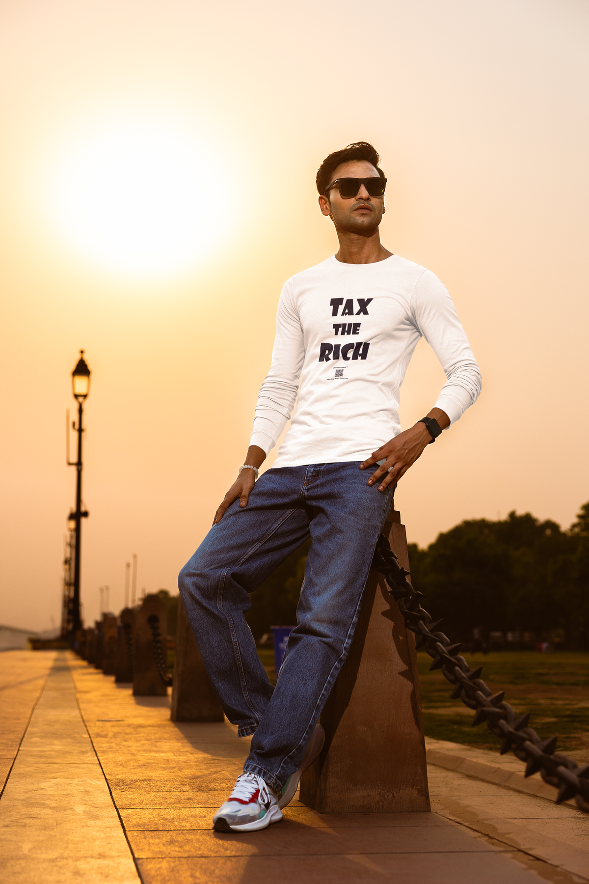 A man wearing a white long-sleeve t-shirt with 'TAX THE RICH' printed in black on the front, blue jeans, and white sneakers, posing outdoors at sunset next to a stone railing.