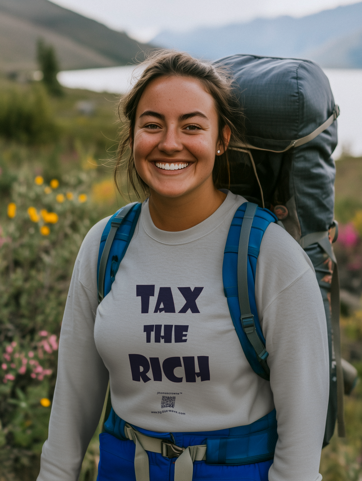 A smiling woman outdoors wearing a large hiking backpack and a light gray long-sleeve shirt with the text 'TAX THE RICH' in dark navy lettering. The background shows a blurred natural landscape with greenery, flowers, and distant hills.
