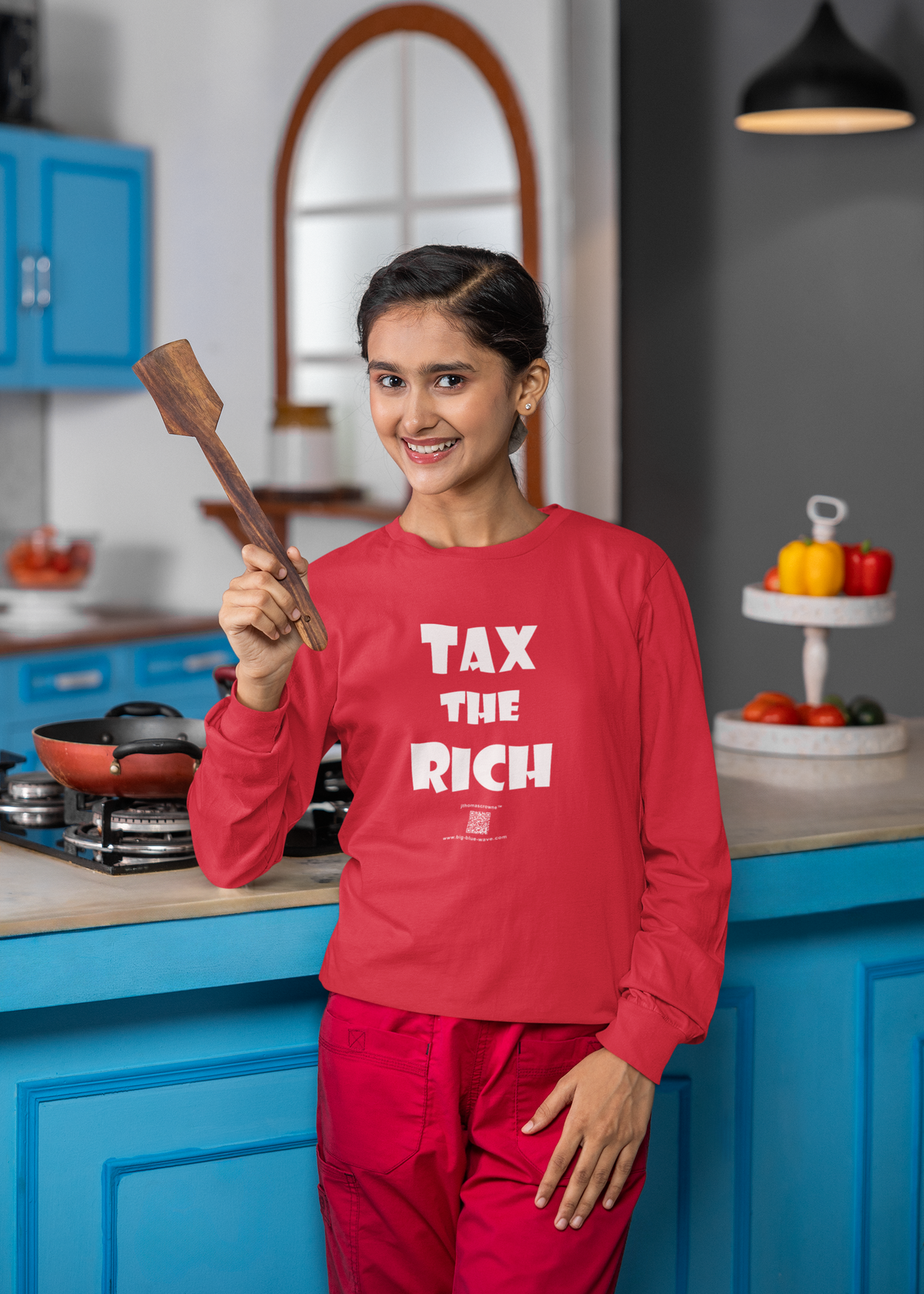 A woman stands in a kitchen wearing a bright red long-sleeve crewneck shirt with the slogan 'TAX THE RICH' printed in bold white letters, paired with matching red pants. She holds a wooden spatula.