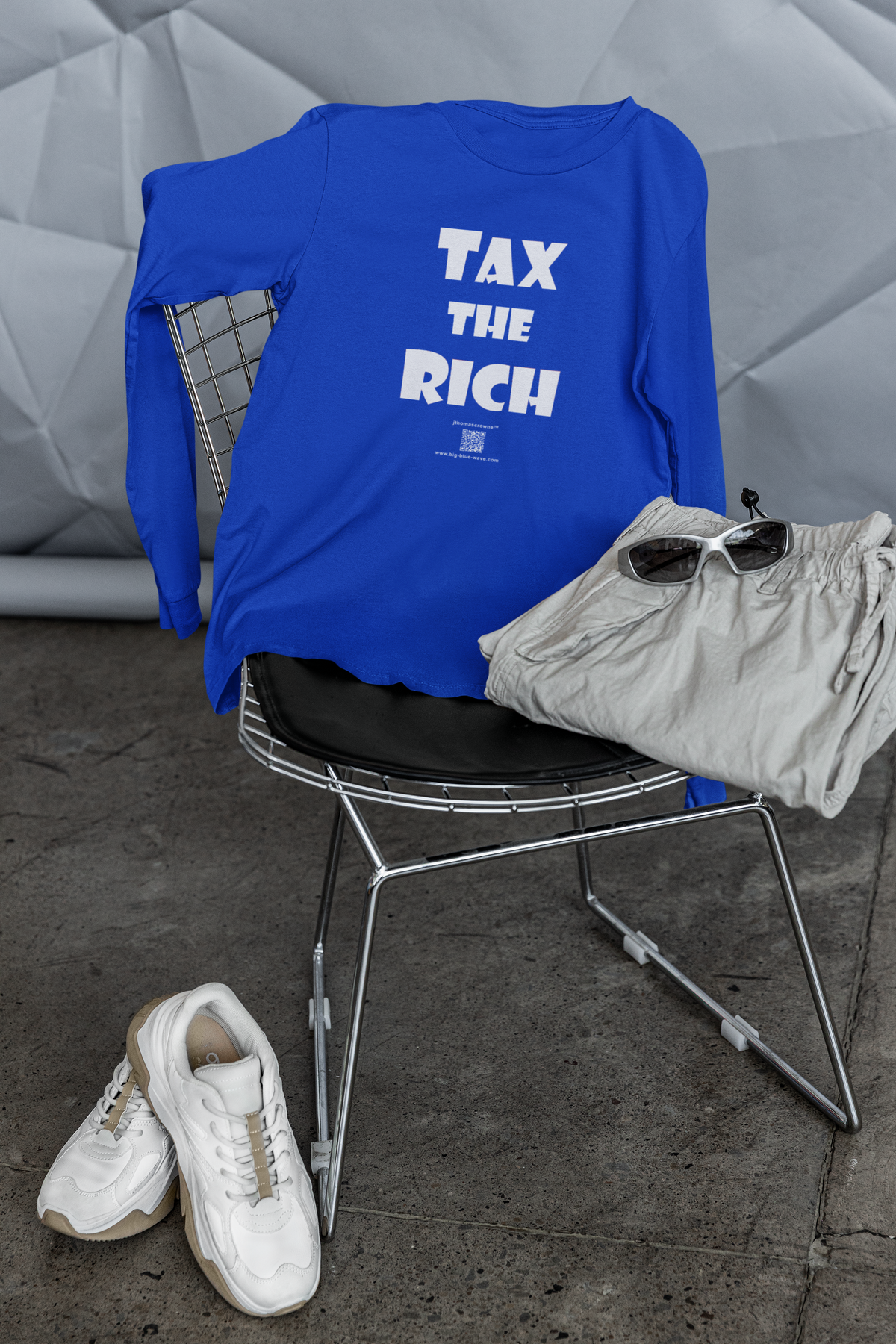 A royal blue long-sleeve t-shirt with the text 'TAX THE RICH' printed in white on the front, draped over a wire chair. A pair of light gray shorts, white sneakers, and sunglasses are styled with the shirt on a metal chair in front of a gray geometric wall.