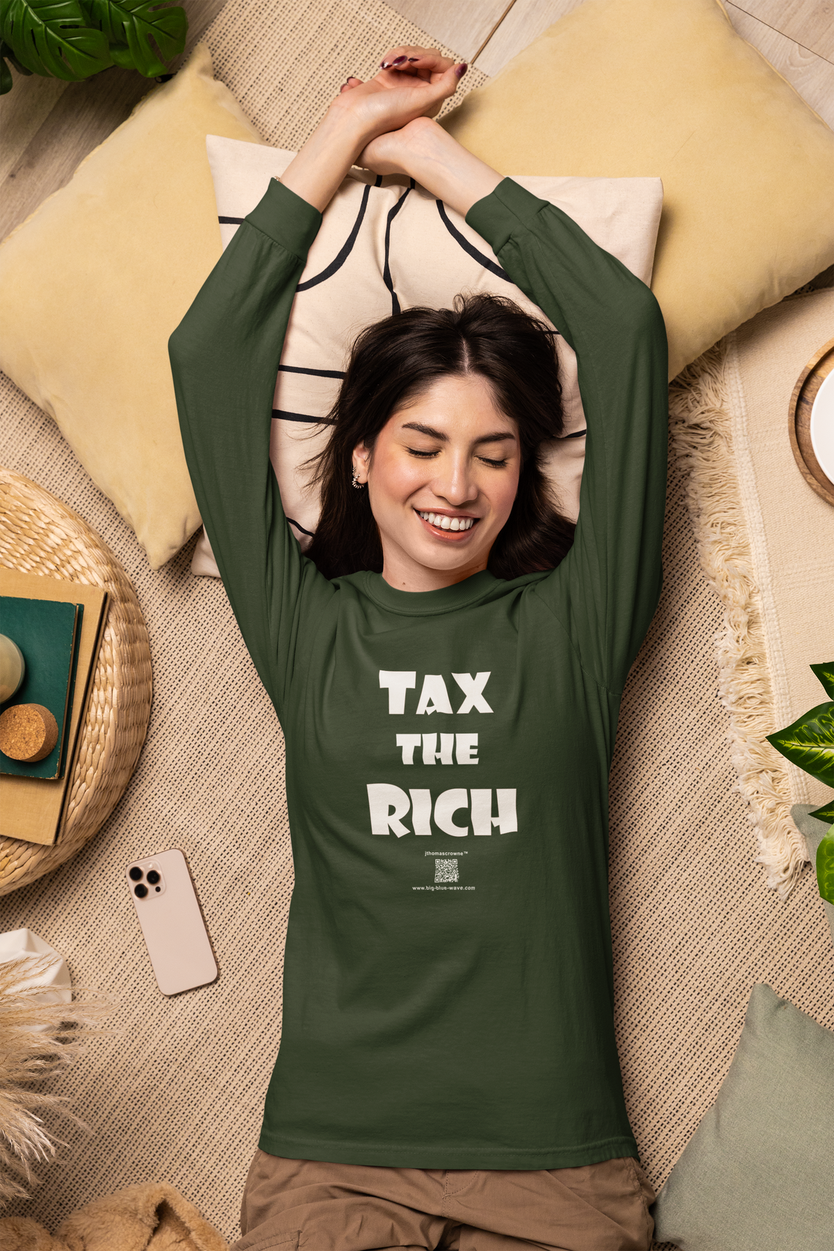 A woman lying on a woven floor with her arms stretched overhead, wearing a dark green long-sleeve t-shirt with the slogan 'TAX THE RICH' printed in bold white letters on the front.