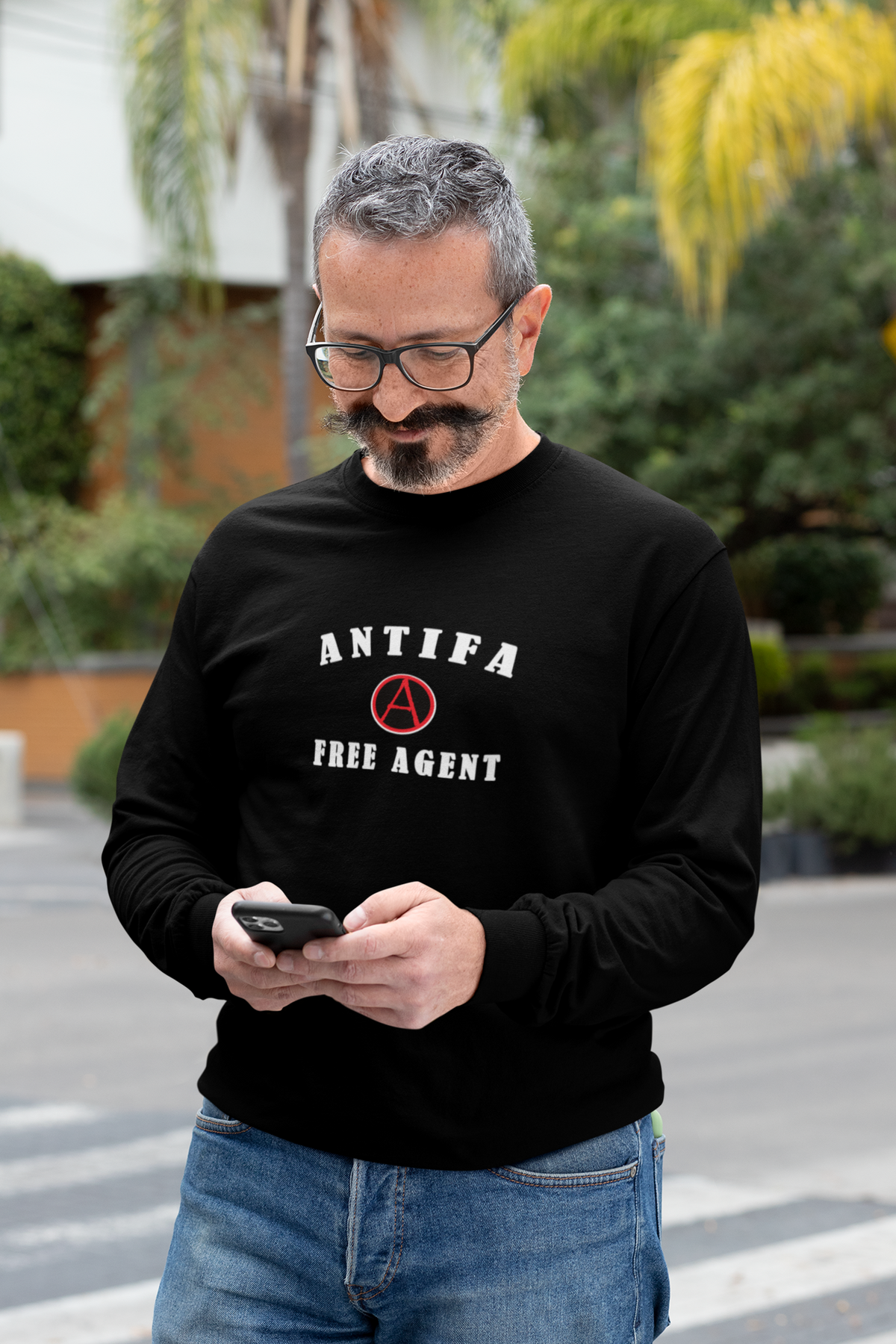 A man wearing a black long-sleeve crewneck sweatshirt with 'ANTIFA FREE AGENT' printed in white and a red anarchist symbol on the chest, paired with blue jeans, standing outdoors and looking at his phone.