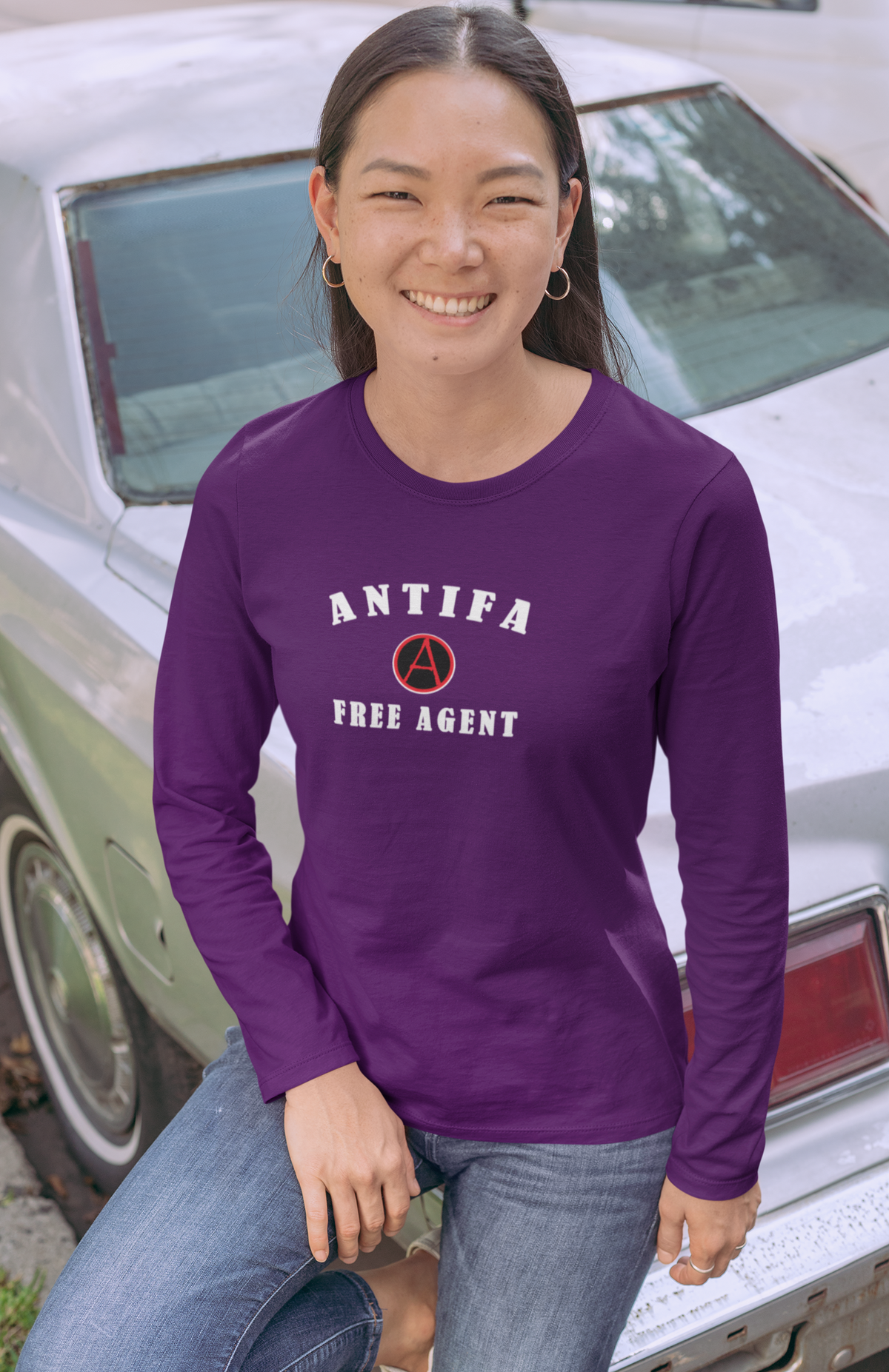 A woman wearing a purple long-sleeve t-shirt with a white graphic that reads 'ANTIFA FREE AGENT' and a red circle-A symbol, posing in front of a vintage car.