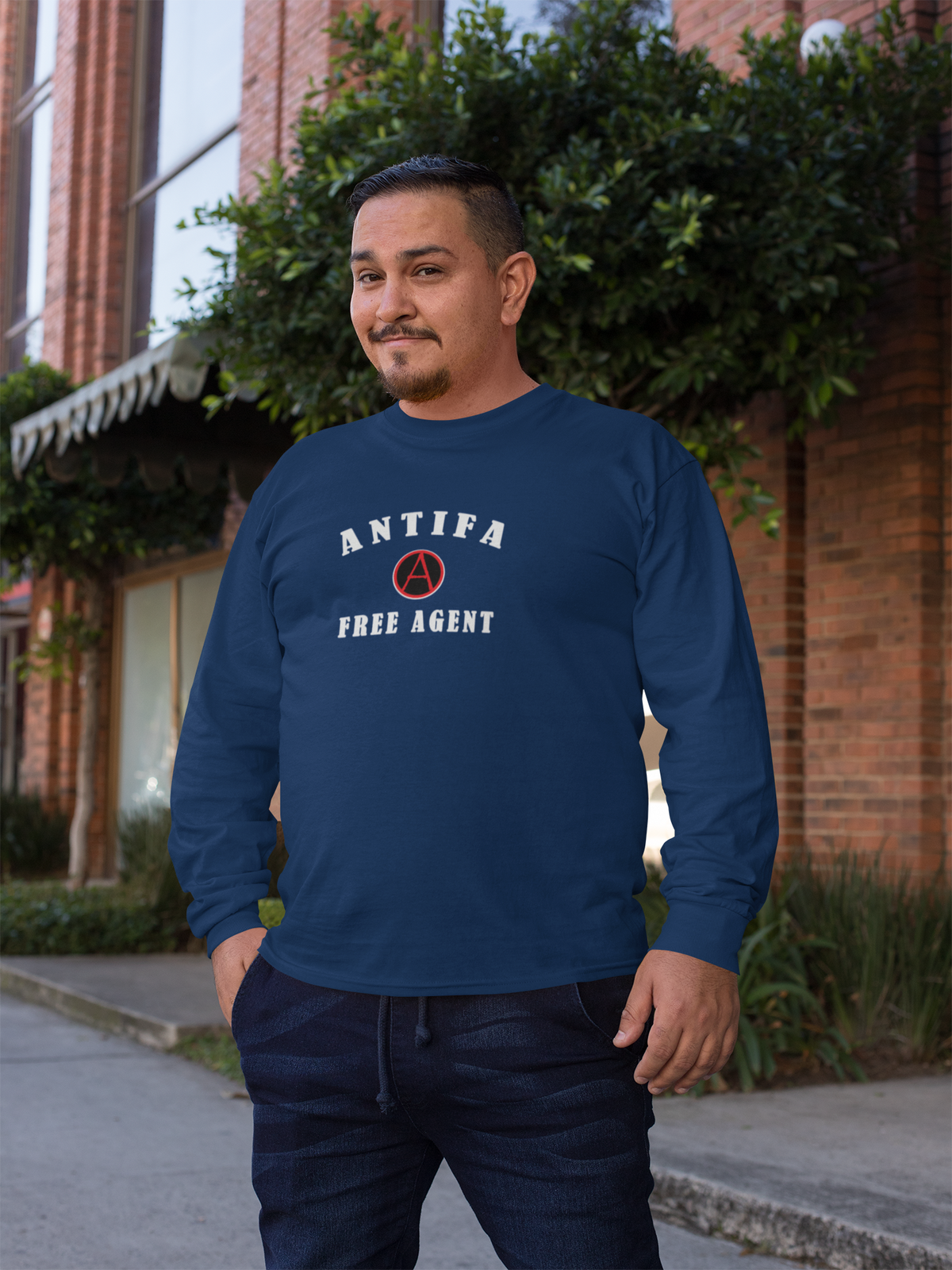 A man wearing a navy blue long-sleeve t-shirt with 'ANTIFA FREE AGENT' and a red-and-white circular emblem printed on the chest, standing outdoors in front of a brick building and greenery.