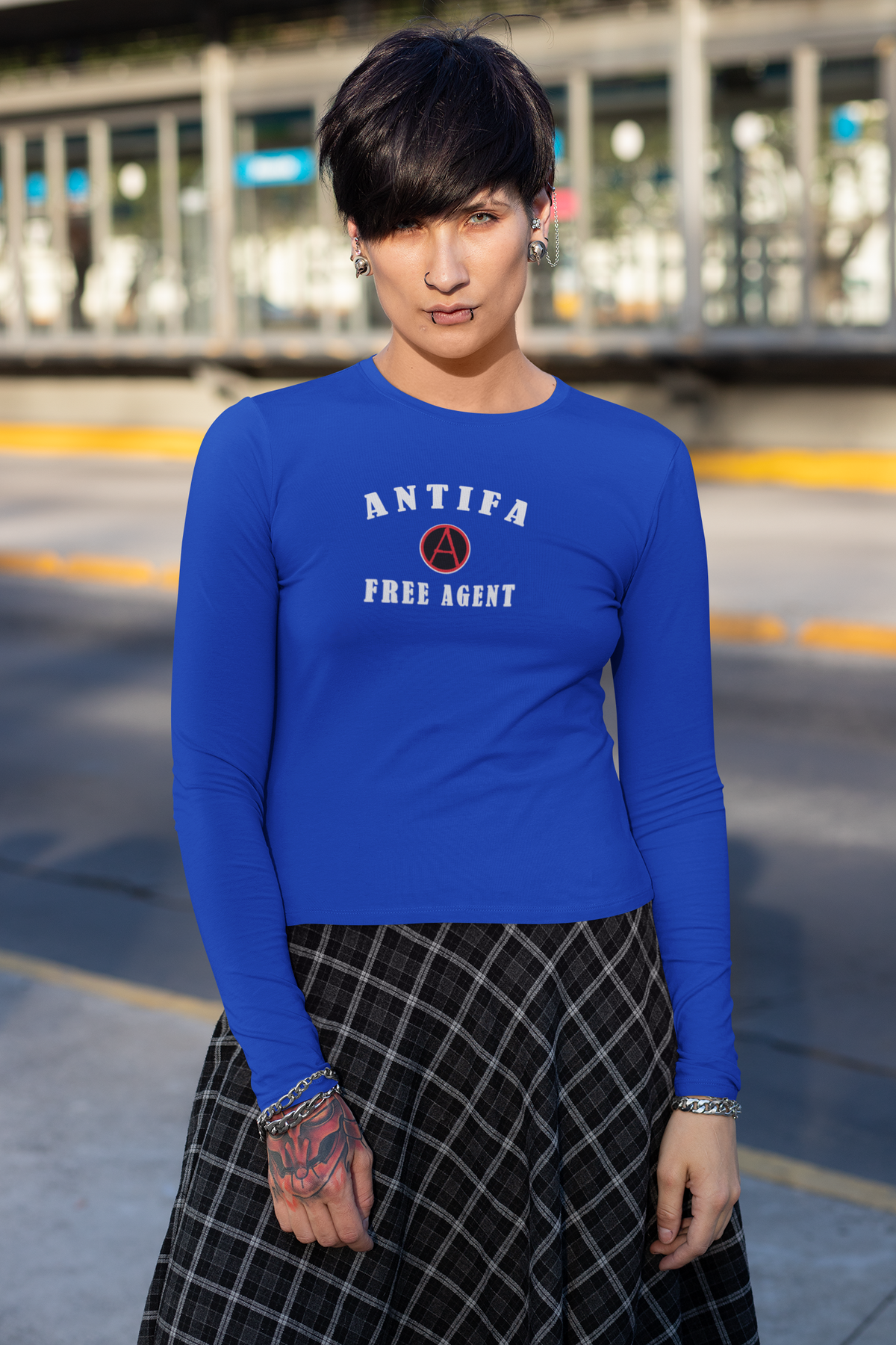 A person wearing a bright blue long-sleeve crewneck shirt with 'ANTIFA FREE AGENT' and a circular A logo printed on the chest, paired with a black and white plaid skirt, standing outdoors in front of a blurred street background.