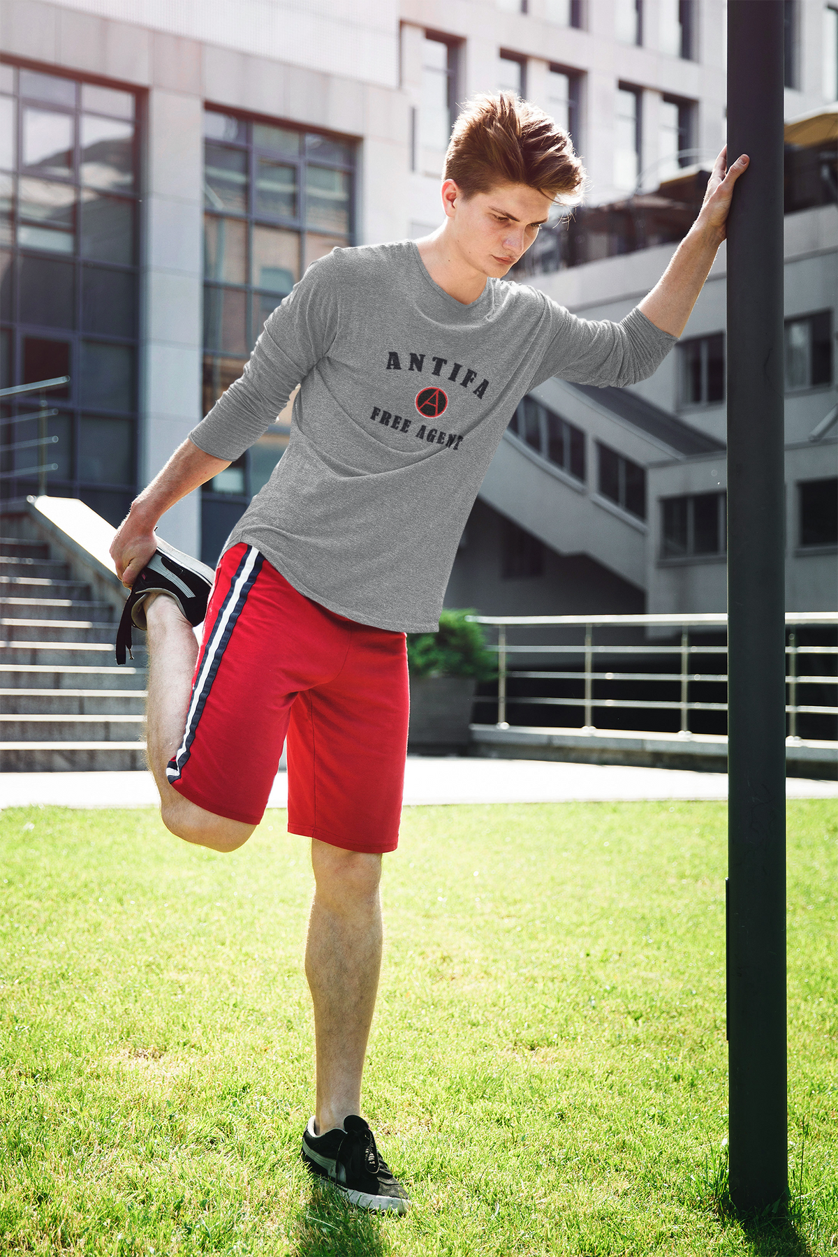 A man in a gray long-sleeve athletic shirt with 'ANTIFA THREE SALUT' printed on the chest and red athletic shorts with a white and blue stripe, stretching his leg outdoors on a grassy lawn with a modern building in the background.