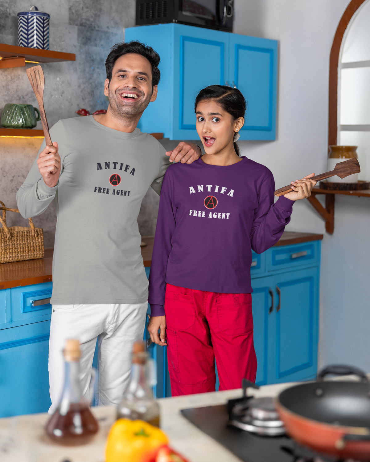 A man and a girl standing in a kitchen, both wearing matching long-sleeve crewneck sweatshirts with 'ANTIFA FREE AGENT' printed on the front. The man wears a light gray sweatshirt with white pants, and the girl wears a purple sweatshirt with red pants.