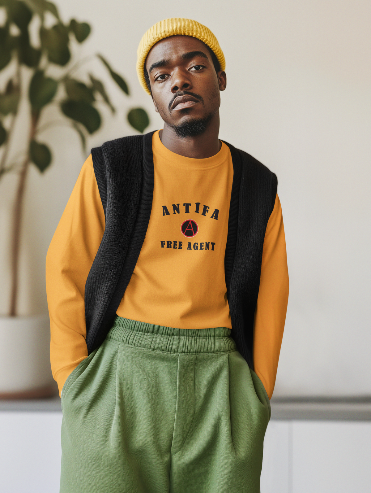 A man wearing a yellow beanie, a yellow crewneck sweatshirt with 'ANTIFA FREE AGENT' text, a black knit vest, and olive green sweatpants, standing indoors against a light wall with a plant in the background.