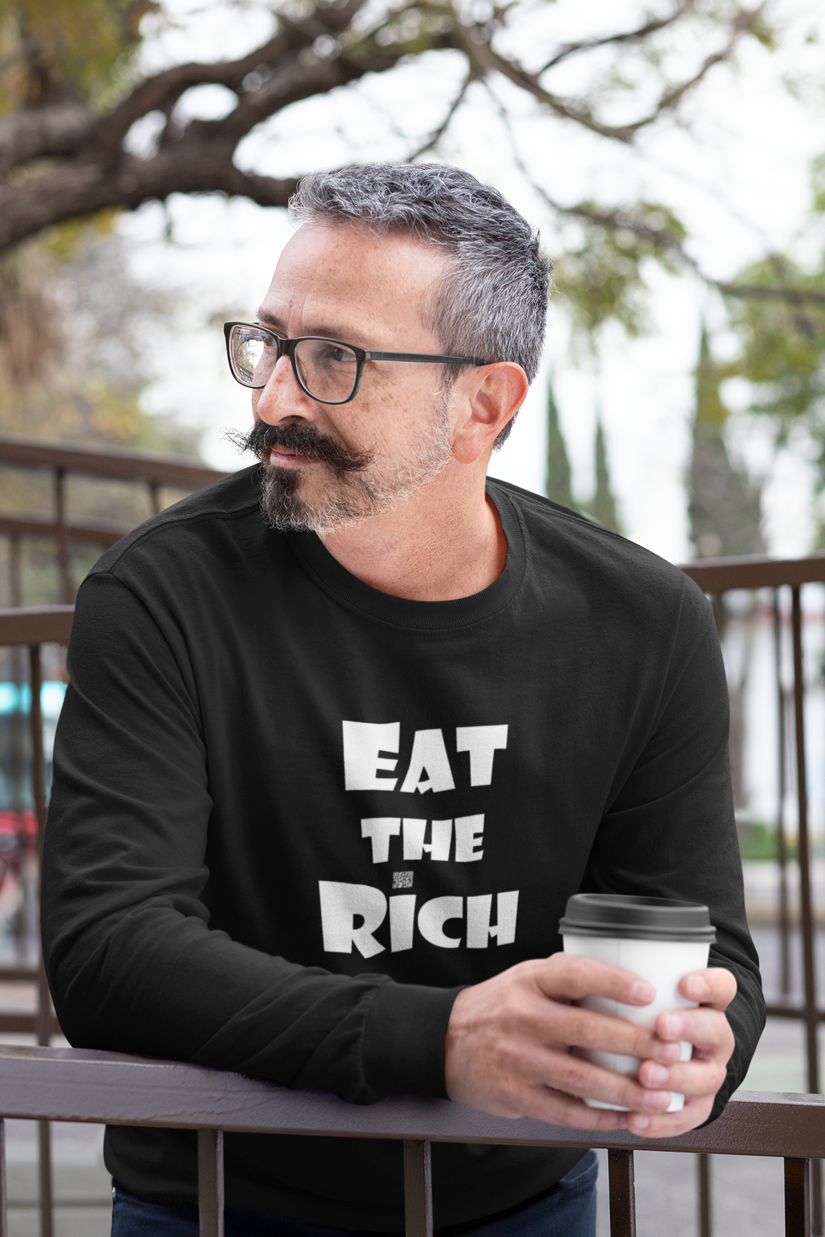 A man with a mustache and glasses wearing a black long-sleeve t-shirt with the text 'EAT THE RICH' printed in white on the front, holding a white paper coffee cup while leaning on a railing.