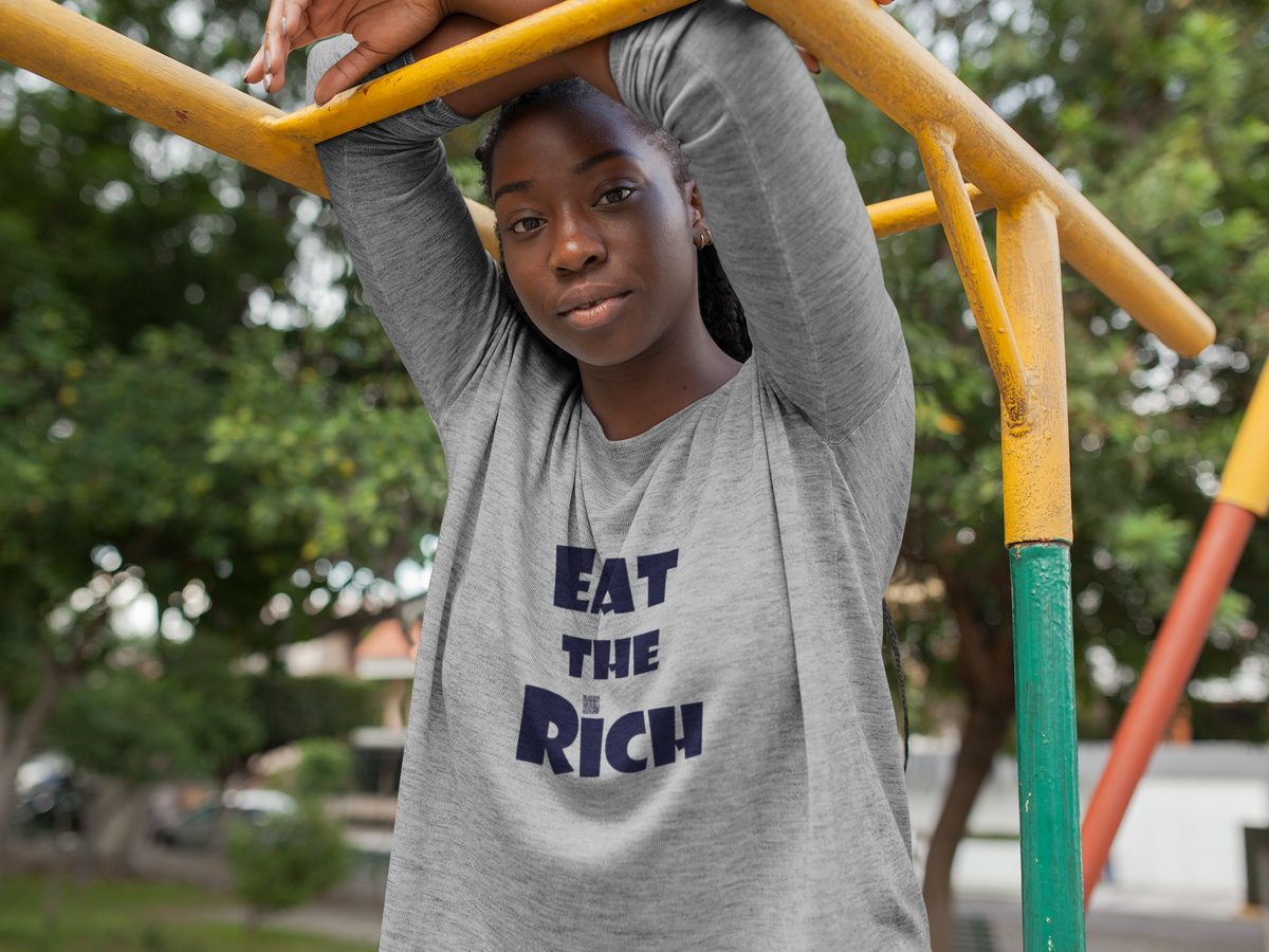 A young woman wearing a heather gray long-sleeve t-shirt with the text 'EAT THE RICH' printed in navy blue on the front, posing on yellow playground monkey bars outdoors.