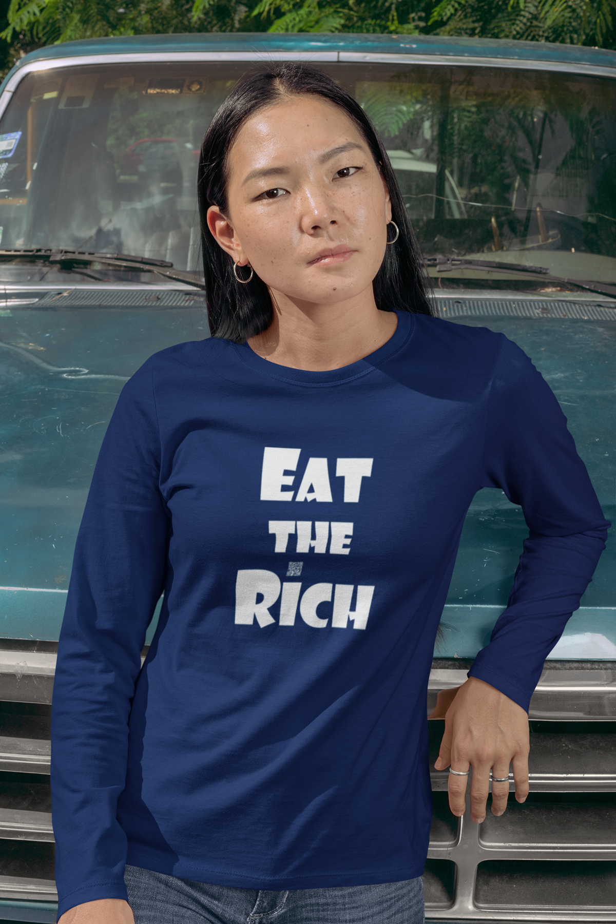 A woman wearing a navy blue long-sleeve t-shirt with the text 'EAT THE RICH' printed in bold white letters on the front, posed in front of a vintage teal truck.