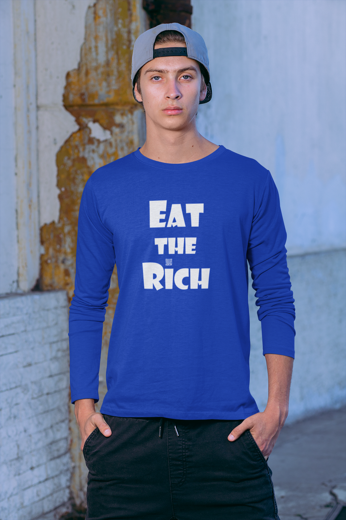 A man wearing a royal blue long-sleeve t-shirt with the slogan 'EAT THE RICH' printed in bold white letters on the front, paired with black pants and a gray baseball cap, standing against a weathered brick wall.