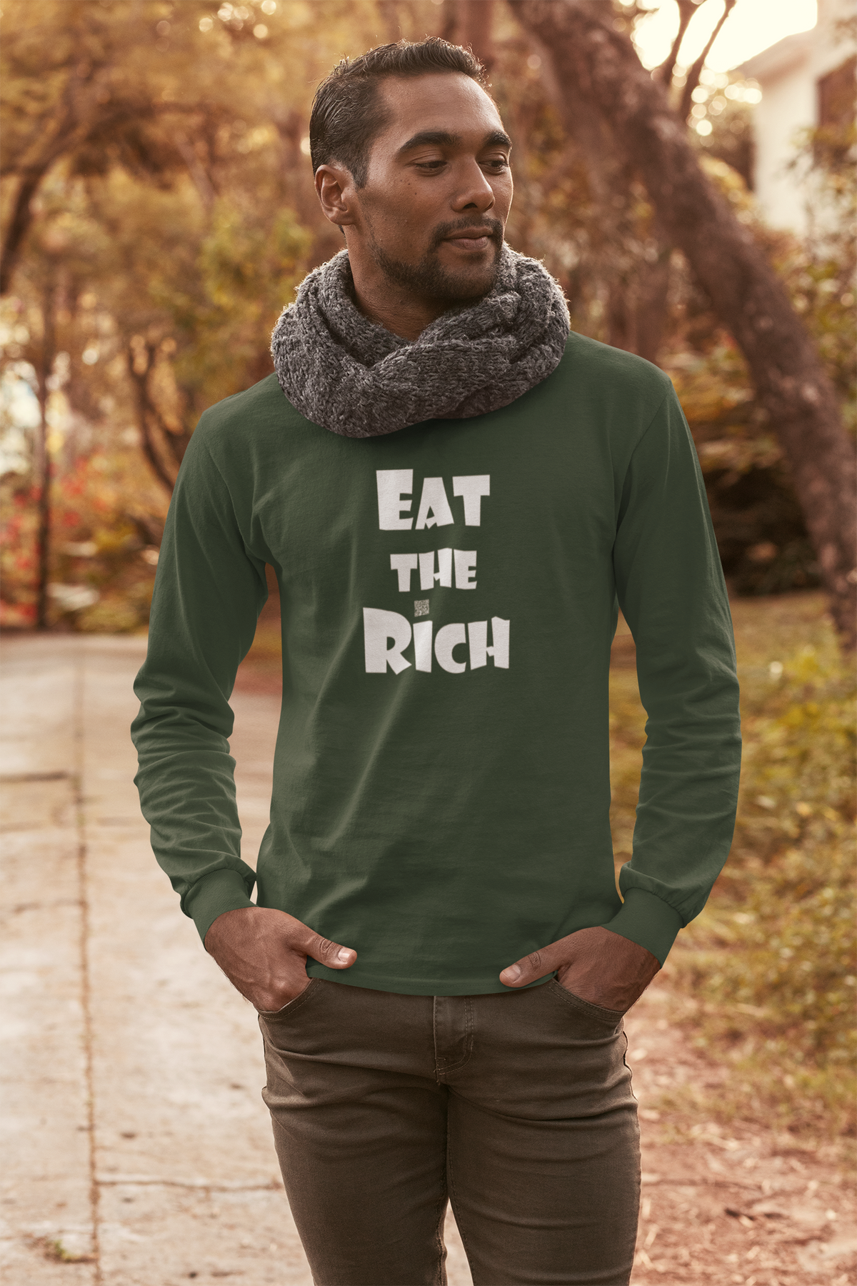 A man wearing a dark green long-sleeve t-shirt with the text 'EAT THE RICH' printed in white on the front, paired with a gray knit scarf and brown pants, standing outdoors on a path with a blurred autumn background.