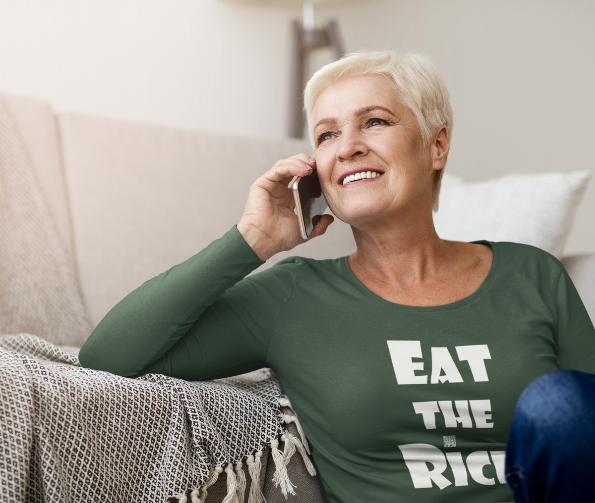 A smiling older woman with short blonde hair sits on a couch, holding a phone to her ear. She is wearing a dark green long-sleeve shirt with the white text 'EAT THE RICH' printed on the front.