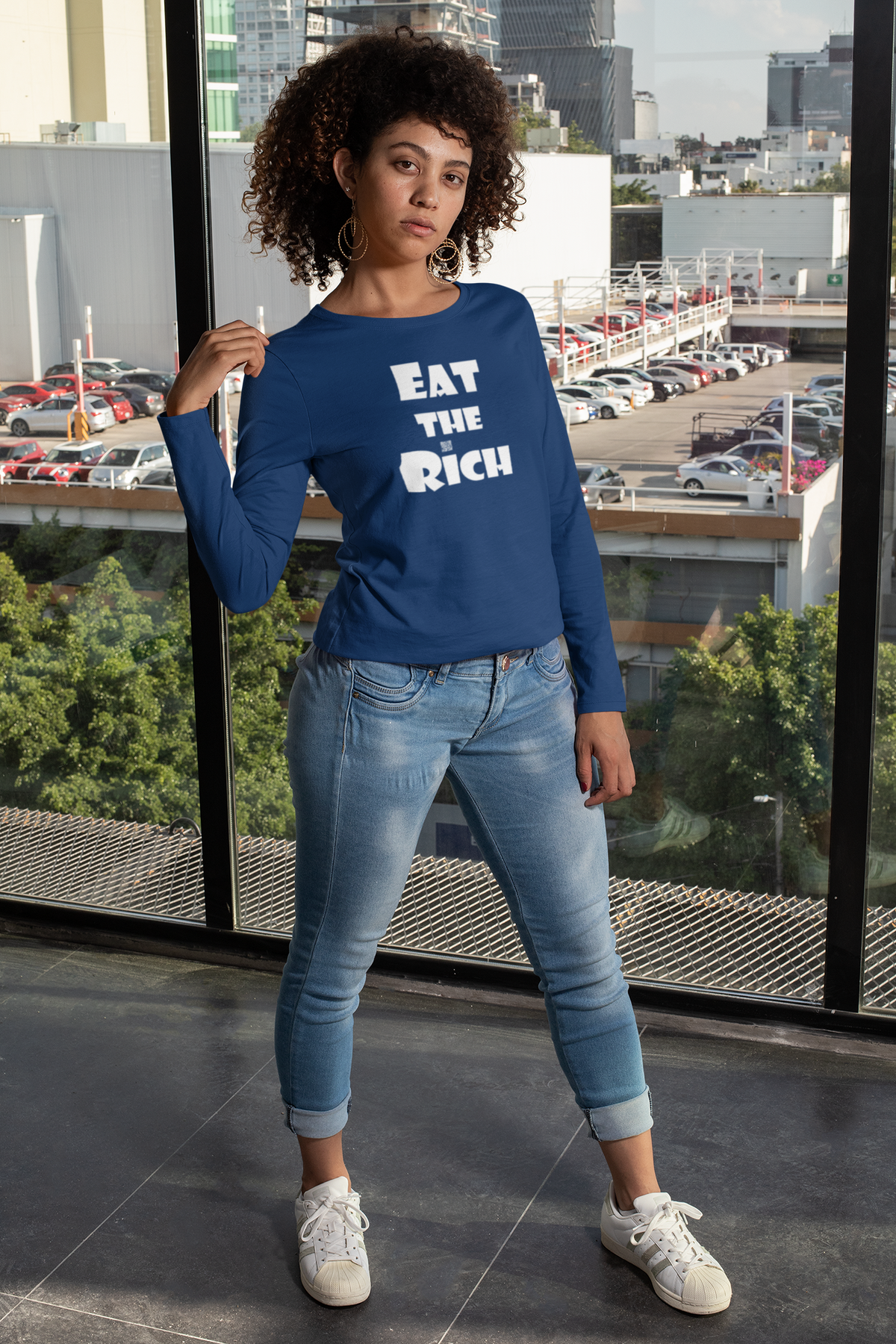 A smiling older woman with short blonde hair sits on a couch, holding a phone to her ear. She is wearing a dark green long-sleeve shirt with the white text 'EAT THE RICH' printed on the front.