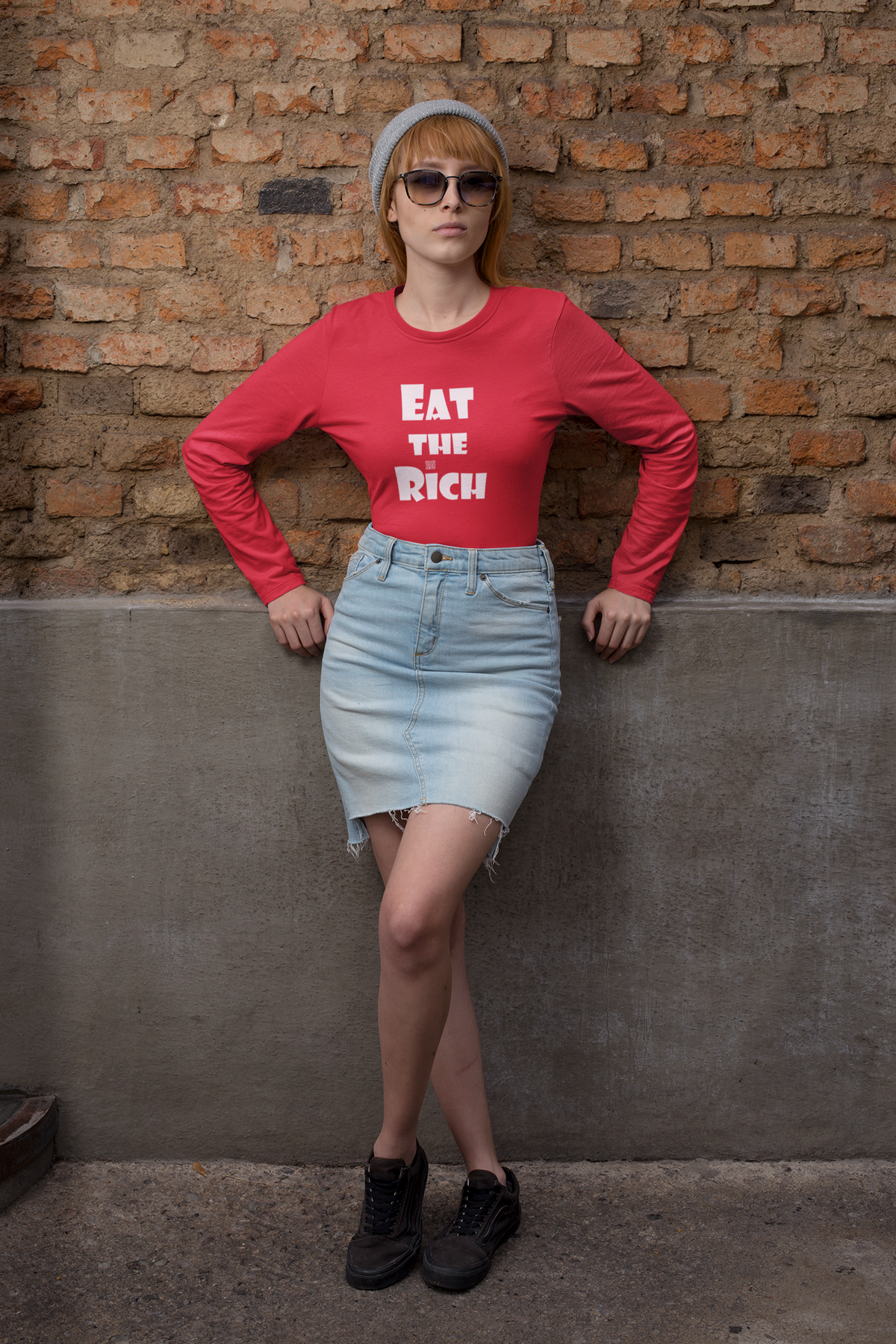 A woman posing against a brick wall wearing a red long-sleeve t-shirt with the text 'EAT THE RICH' in white, a light blue denim mini skirt, black sneakers, and a gray beanie.
