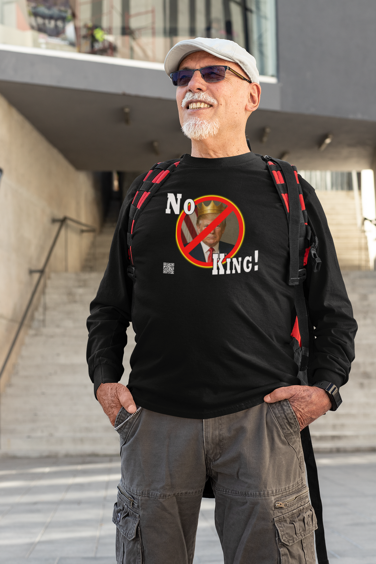 An older man wearing a black graphic t-shirt with a 'No King!' design, gray cargo pants, and a white cap, standing outdoors in front of a modern building.